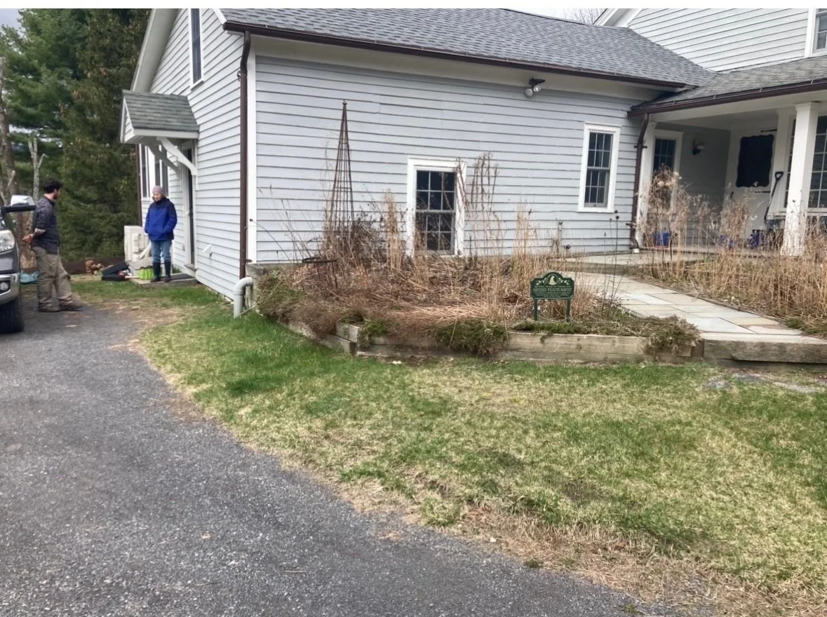 Two people standing outside a house near a driveway with a parked vehicle. The house has light-colored siding, a porch, and a small garden bed with dried plants and a sign that reads 'Ward Lake Historical Society'.