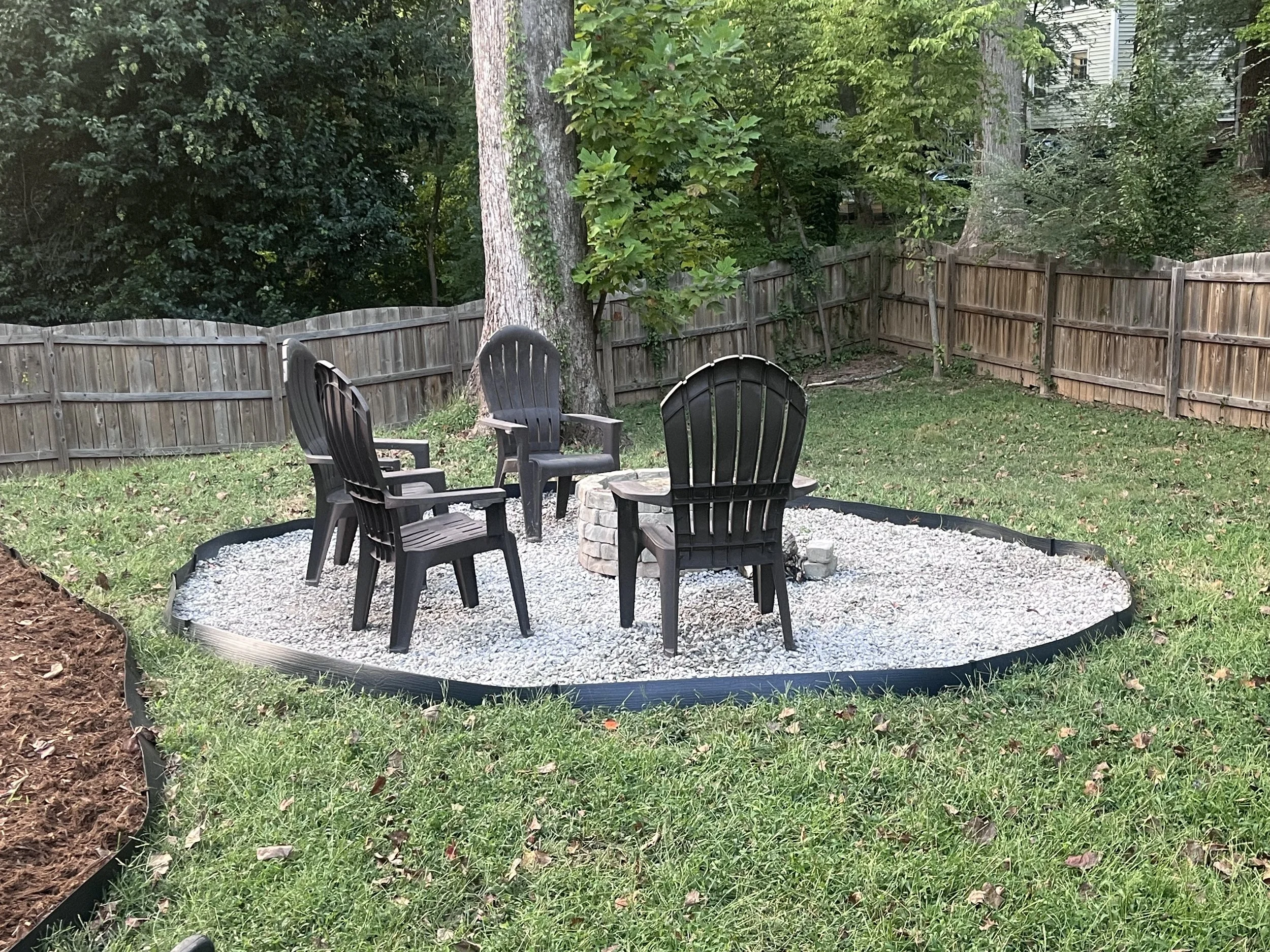 A backyard scene with four black plastic Adirondack chairs arranged around a fire pit on a gravel surface, surrounded by a wooden fence and lush green trees.