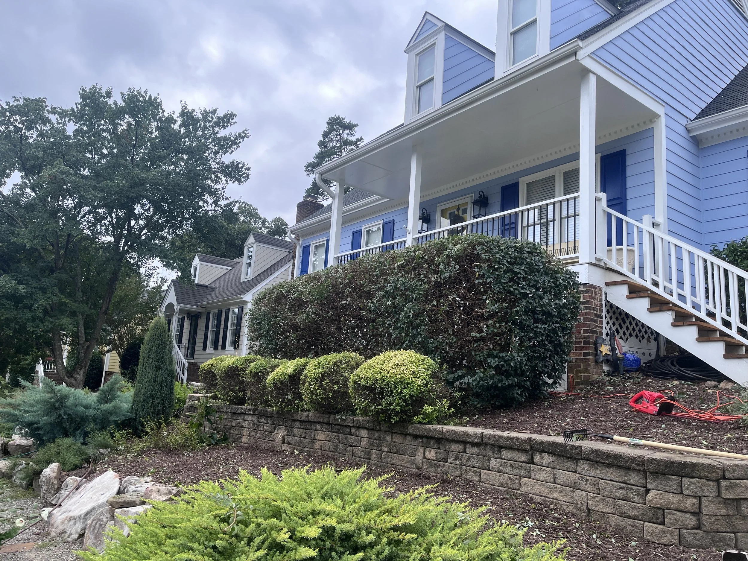 A blue house with white trim and a front porch, surrounded by green bushes and trees, with some garden tools and hoses on the ground.