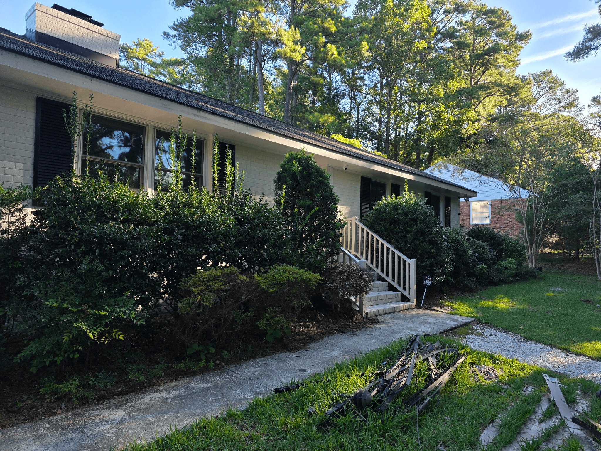 View of a residential house with a porch, stairs, and a garden area in front, surrounded by trees and greenery, under a blue sky.