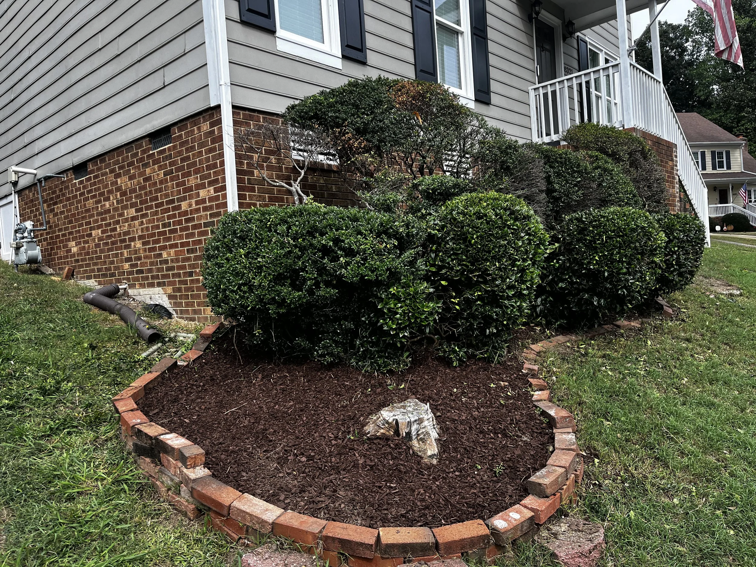A landscaped front yard in front of a house with gray siding and brick foundation. There is a rectangular flower bed outlined with bricks, filled with mulch, and a large decorative rock in the center. Bushes are planted along the house and the yard has green grass.