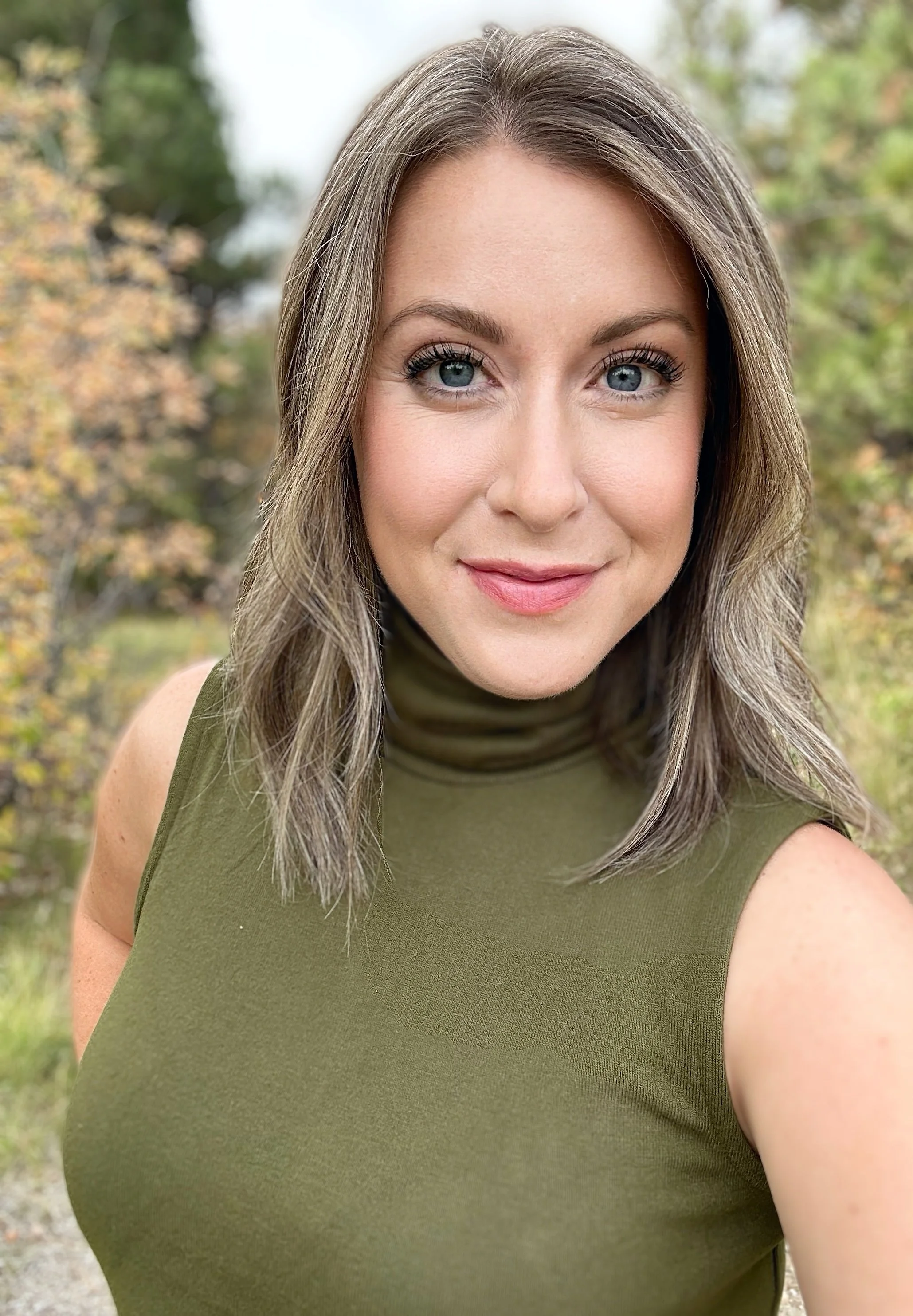 Close-up of Emilie Ritter, a woman with light brown hair and blue eyes, smiling outdoors, wearing an olive green sleeveless top, with a blurred autumnal background.