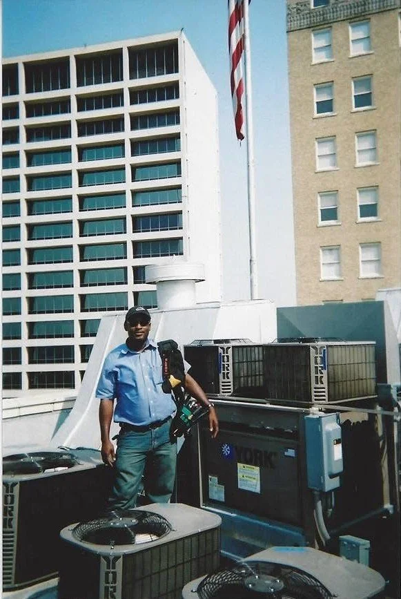A man in a blue shirt and jeans standing on a building rooftop next to air conditioning units with city buildings in the background.