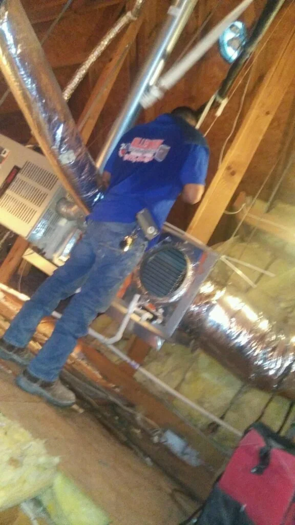 A worker in blue overalls fixing or inspecting an HVAC system in an attic with exposed wooden beams and silver ductwork.