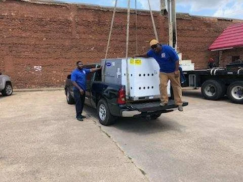 Two men loading a large white industrial air conditioning unit into the back of a pickup truck in an outdoor parking lot.