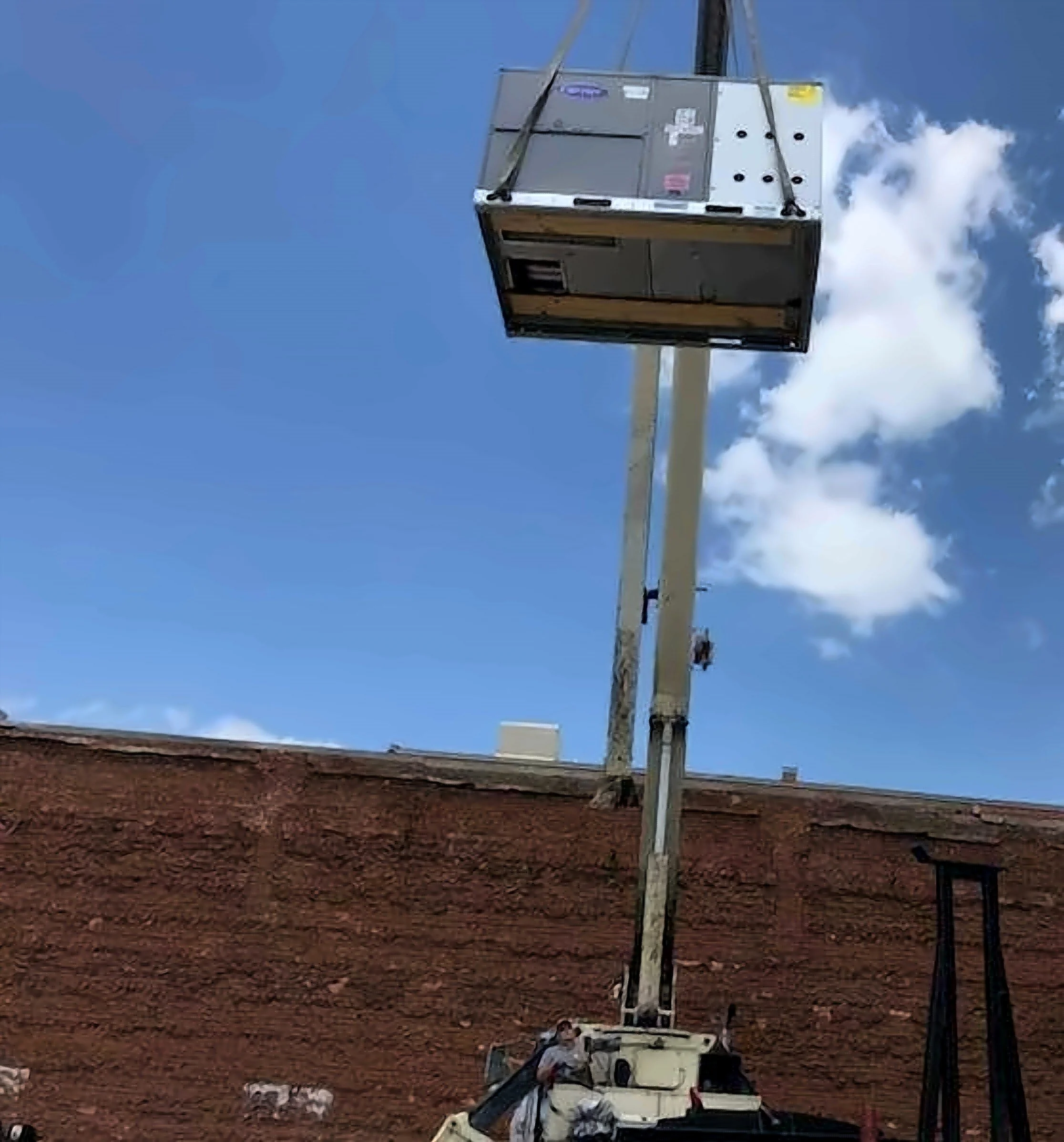 A large piece of construction or industrial equipment mounted on a utility vehicle is shown in a dirt field under a blue sky with some clouds. The equipment appears to be a tall, metallic structure or lifting device.