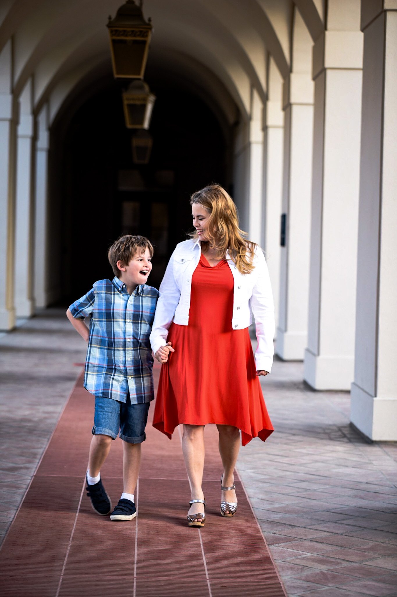 A woman in a red dress and white jacket holding hands with a young boy in a plaid shirt and shorts, walking through a covered outdoor walkway with stone columns and hanging lanterns.