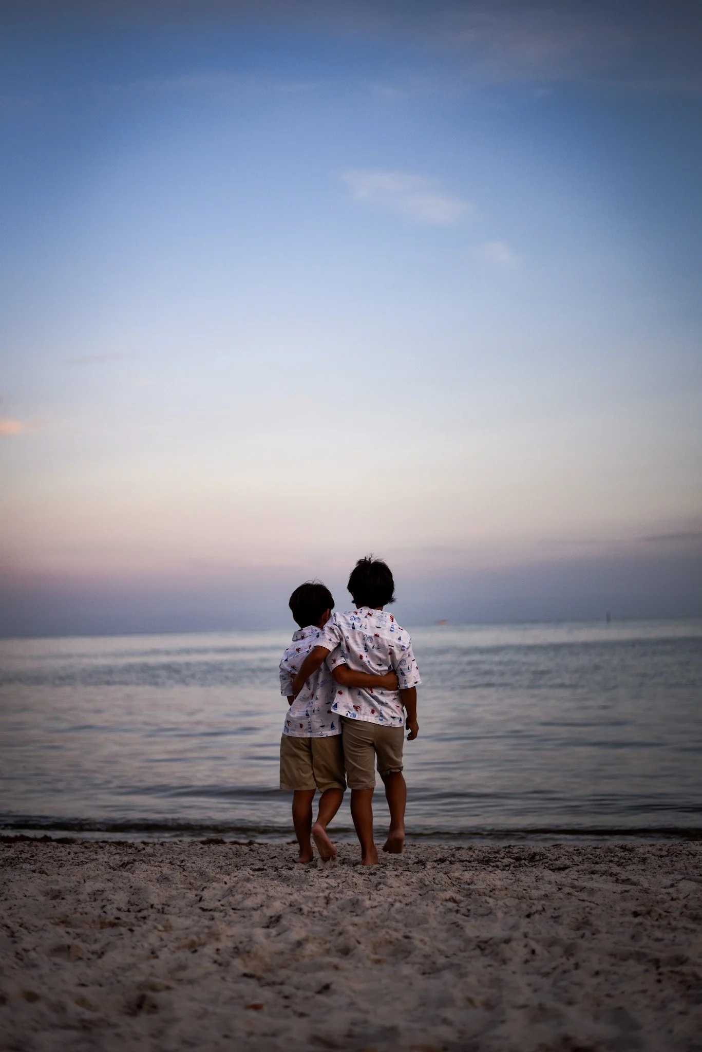Two children hugging on the beach during sunset, facing the ocean with a cloudy sky.