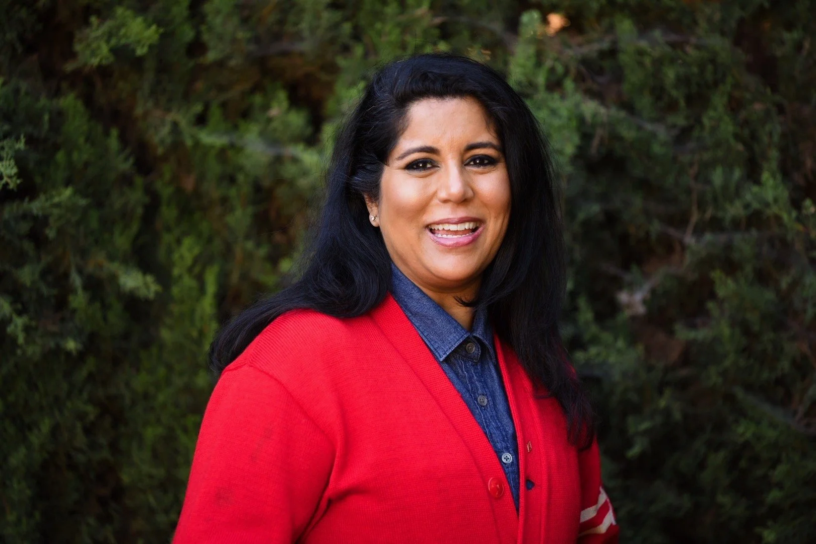 A woman with black hair, wearing a red sweater over a denim shirt, smiling outdoors with green foliage in the background.