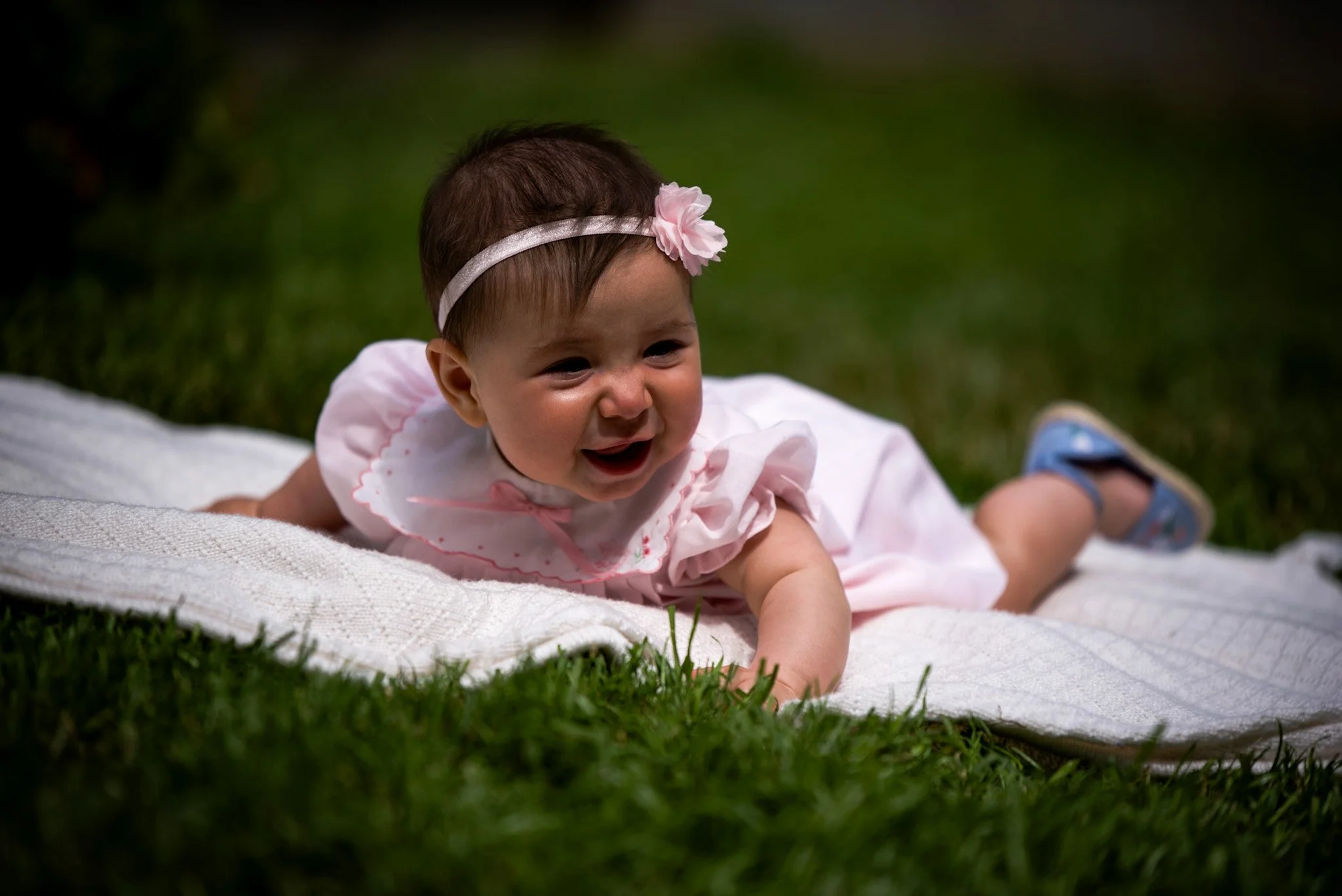 A young baby girl wearing a pink headband with a flower, crawling on a blanket outdoors on green grass.