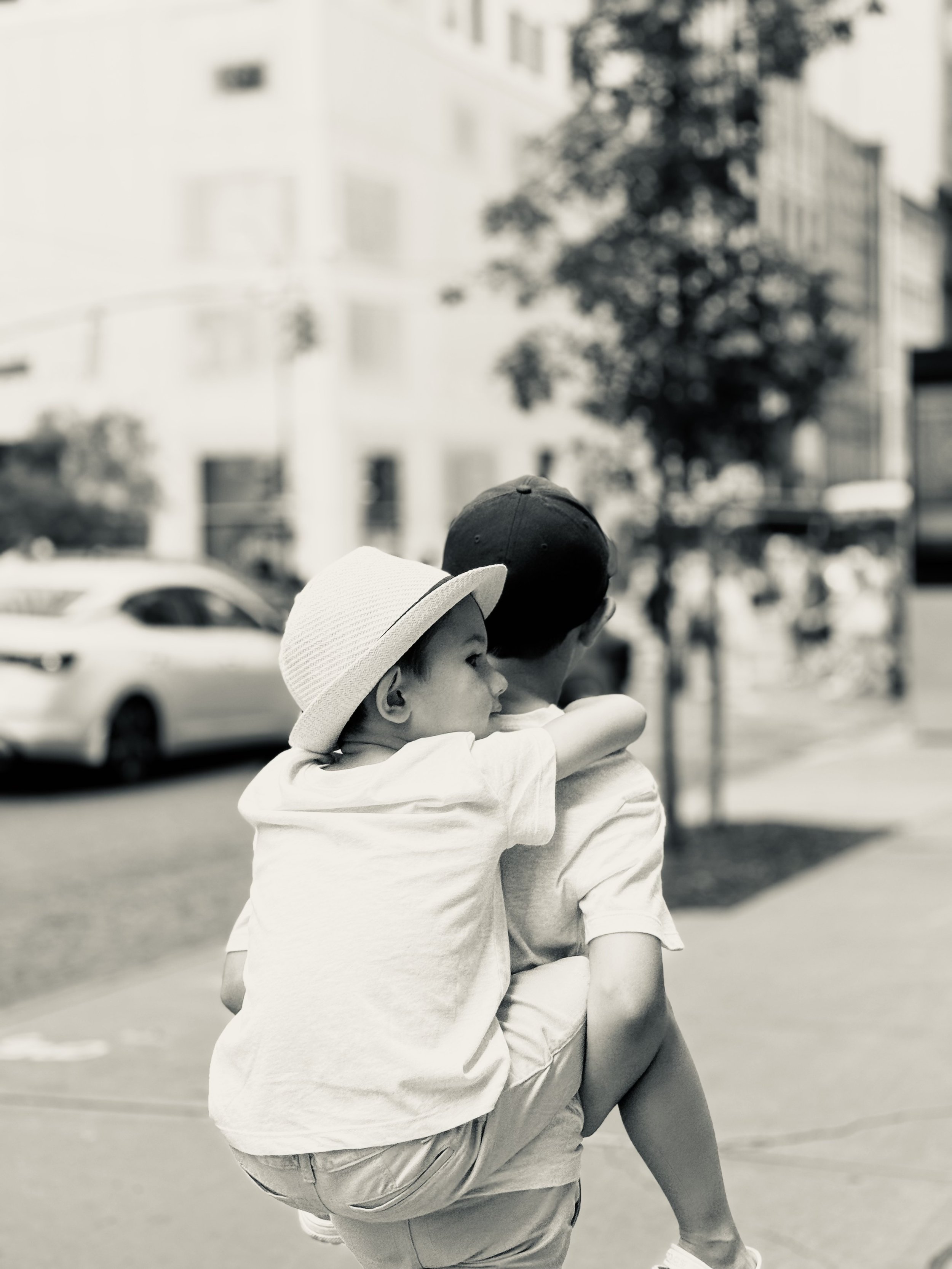 Two young children, a girl and a boy, embrace each other in a hug on a city street. The girl is wearing a white sunhat and the boy a baseball cap. The background features parked cars and trees, suggesting a sunny day.