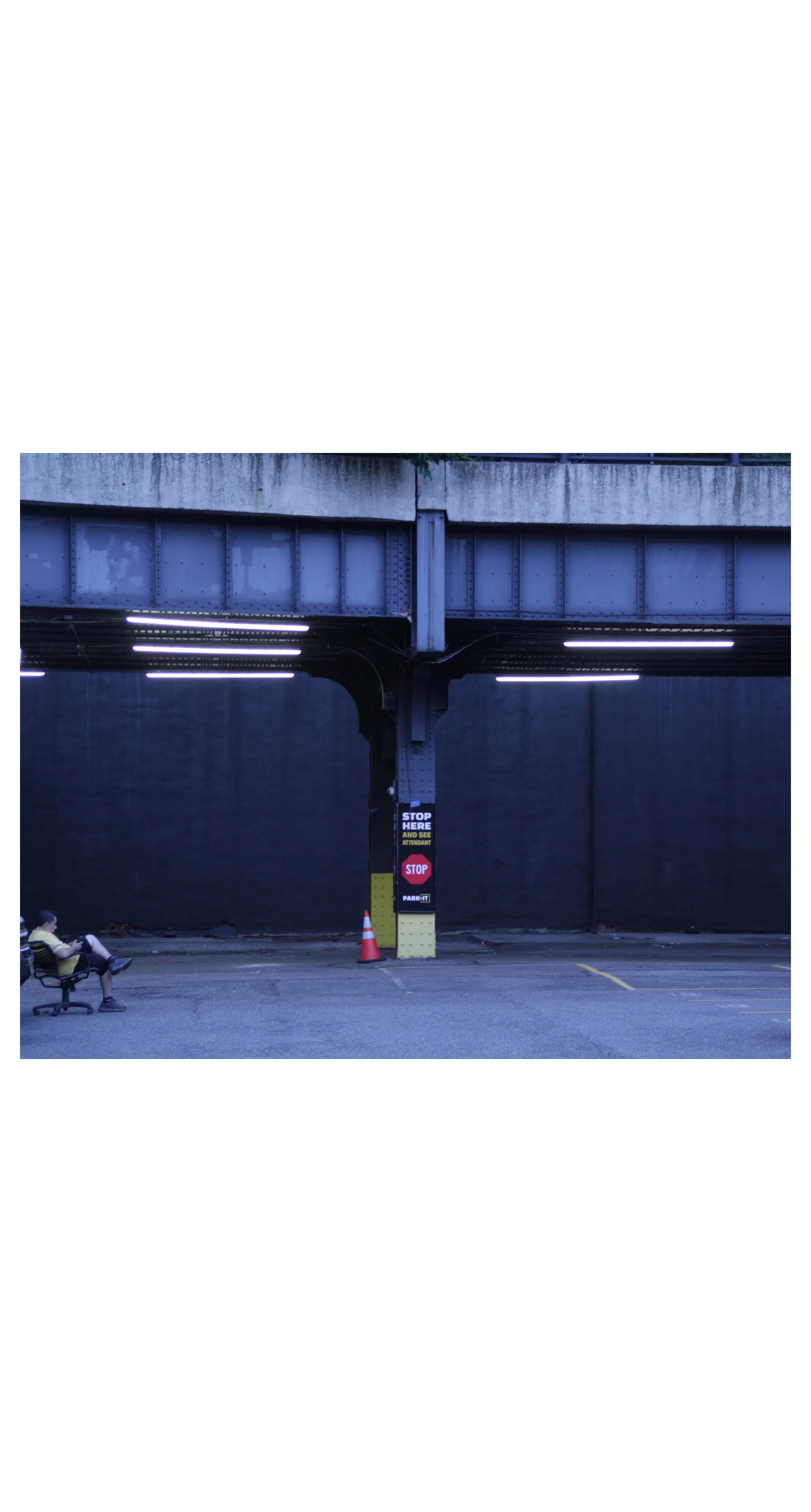 Underpass with a person sitting on a bench to the left, a traffic cone, and parking signs on a pillar under the bridge.