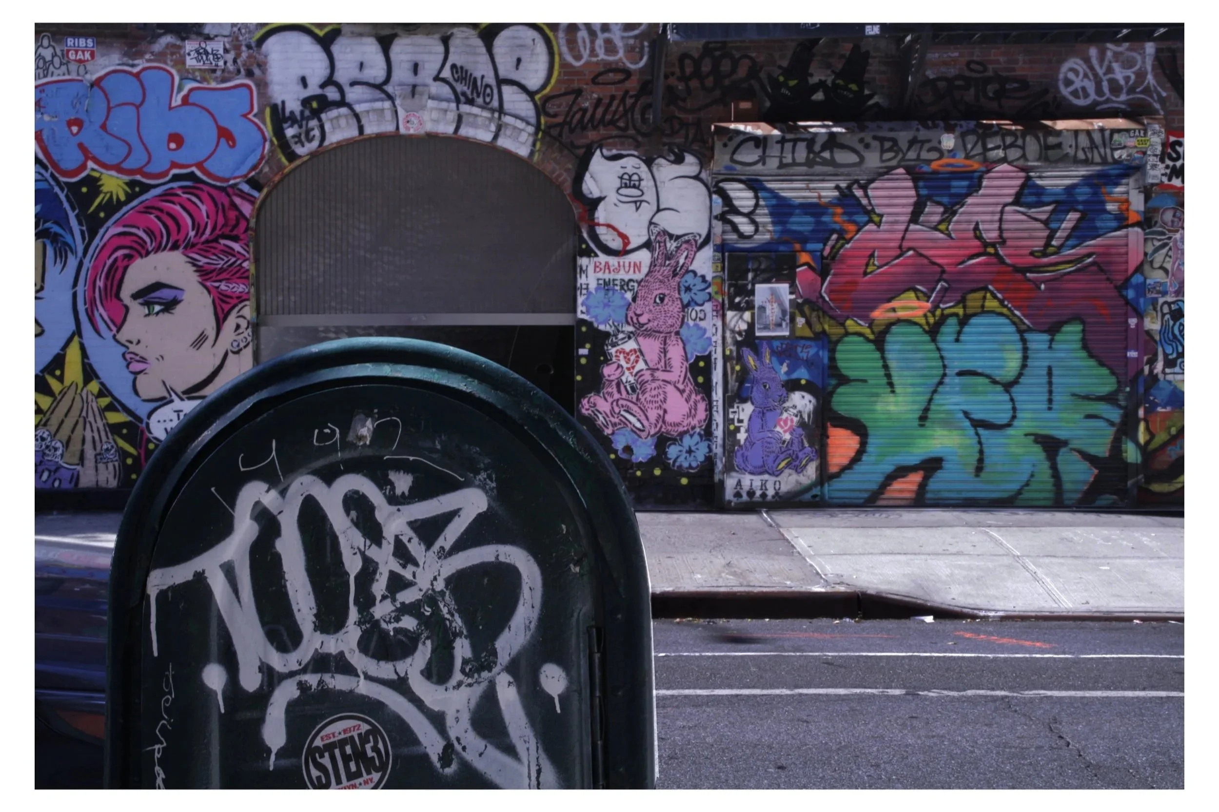 Street art graffiti murals on a brick wall, featuring vibrant characters including a woman with pink hair, a rabbit, and other stylized designs. In the foreground, there is a black round object with white graffiti on it.