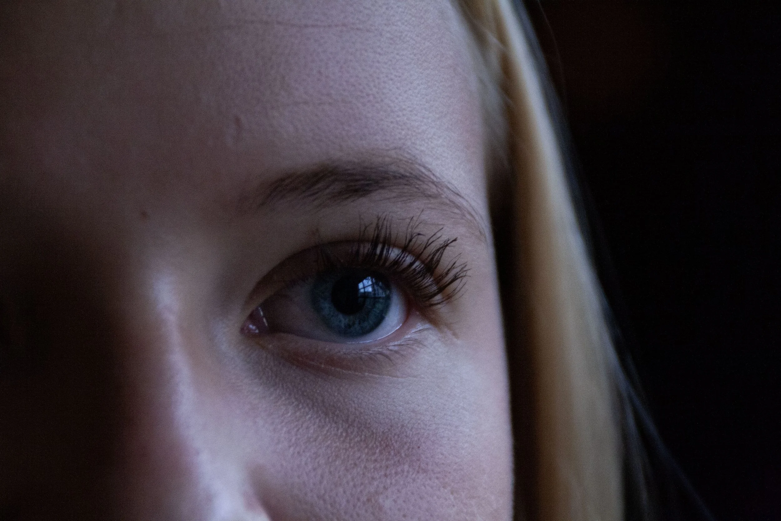 Close-up of a person's face showing one eye with long eyelashes, light skin, closed eyelid, and eye makeup, with soft lighting and dark background.