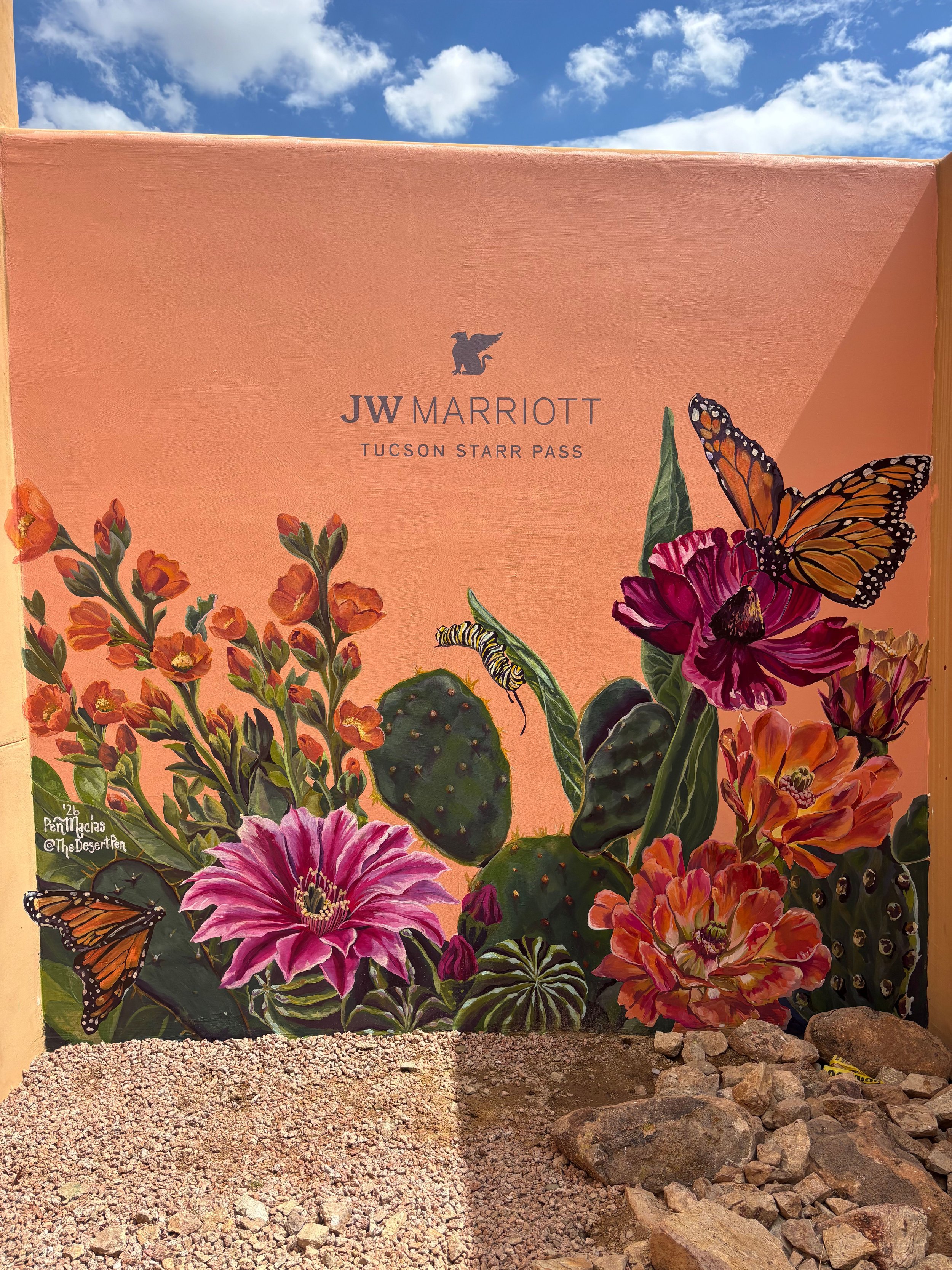 Brightly colored mural with flowers, butterflies, and cacti on an orange background, at the JW Marriott Tucson Starr Pass hotel, with a partly cloudy sky above.