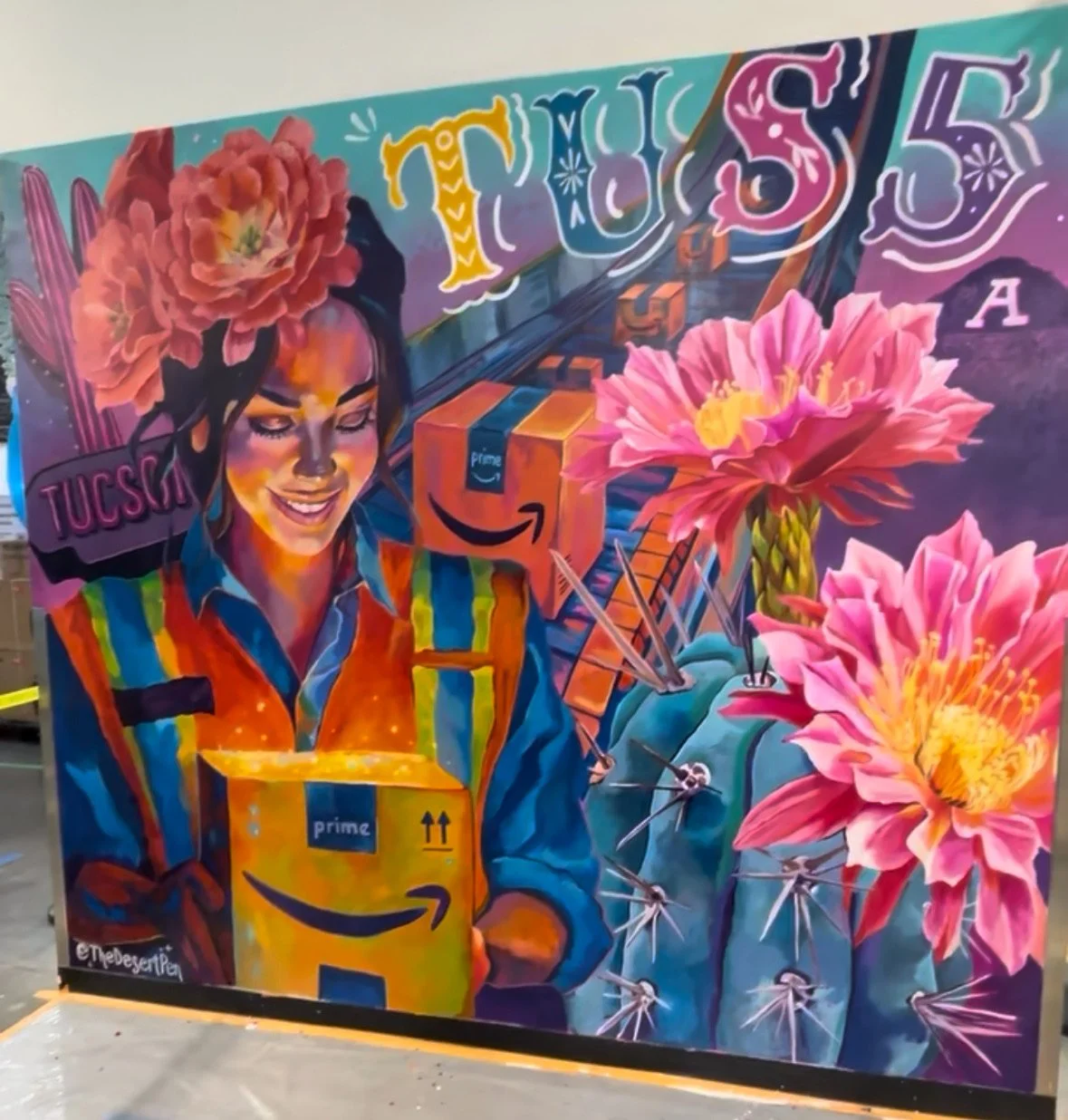 Colorful mural depicting a woman with flowers in her hair holding an Amazon Prime package, with large pink flowers and stylized text including 'TUCSON' and 'TUC'S' in the background.