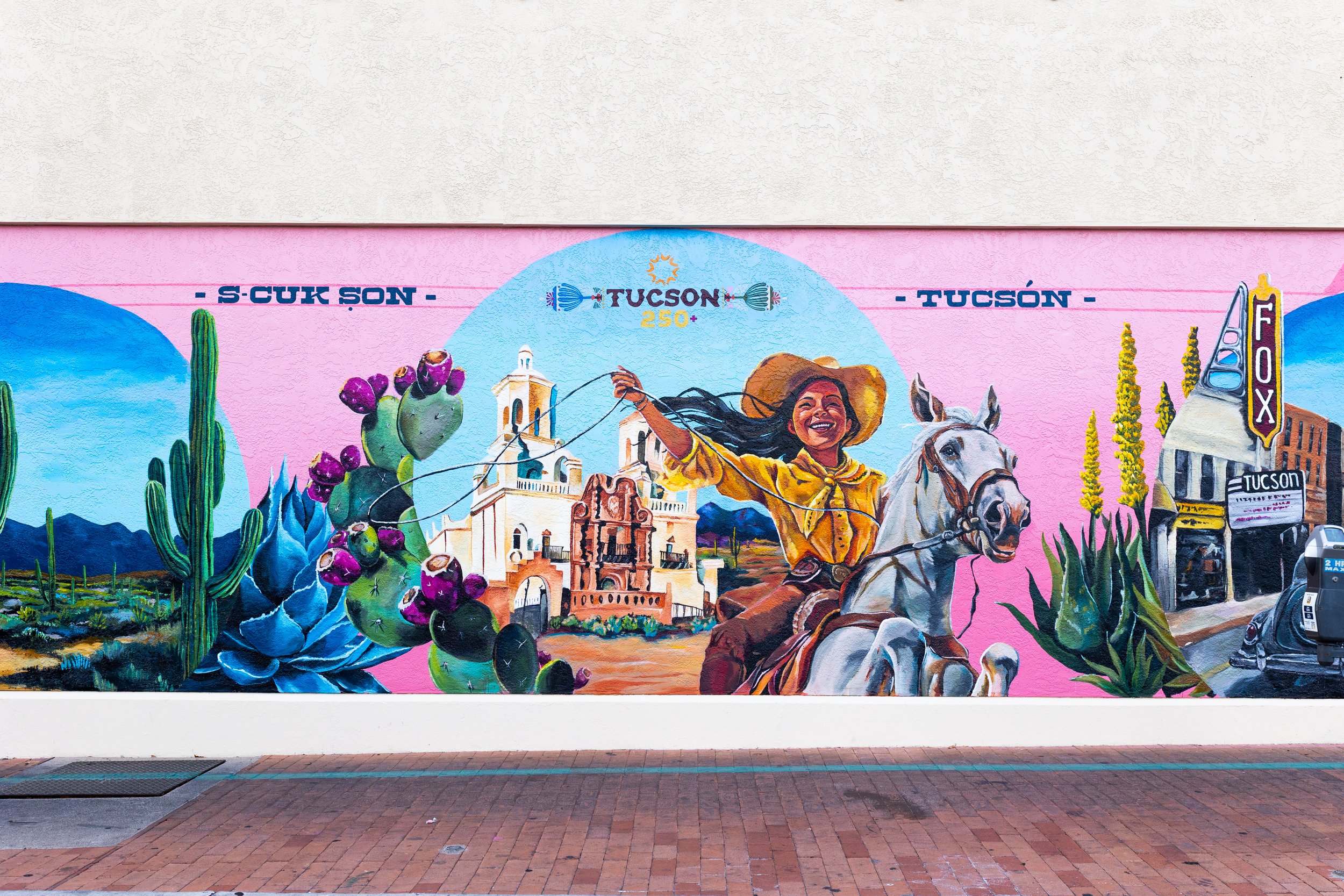 Colorful mural depicting Tucson, Arizona, with desert plants, a smiling woman on horseback, historic buildings, mountains, and signs for Tucson and Foxtown. The mural has a pink background.