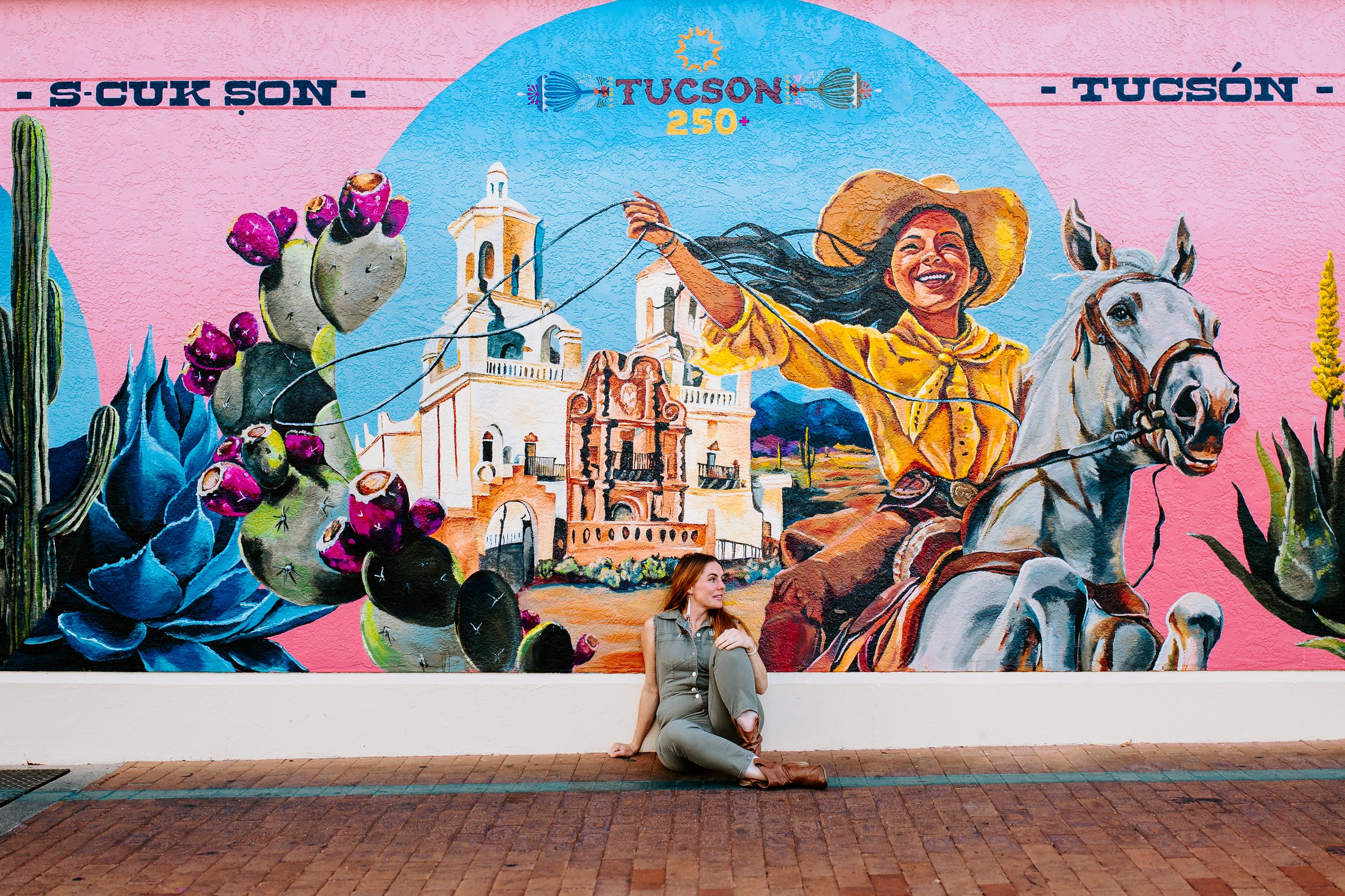 A woman sitting on a brick sidewalk leaning against a white wall in front of a colorful mural. The mural features a smiling woman with long dark hair, a white horse, cacti, and buildings with mountains in the background. The mural also contains the words 'S-CUK SON' and 'TUCSON 250+'.