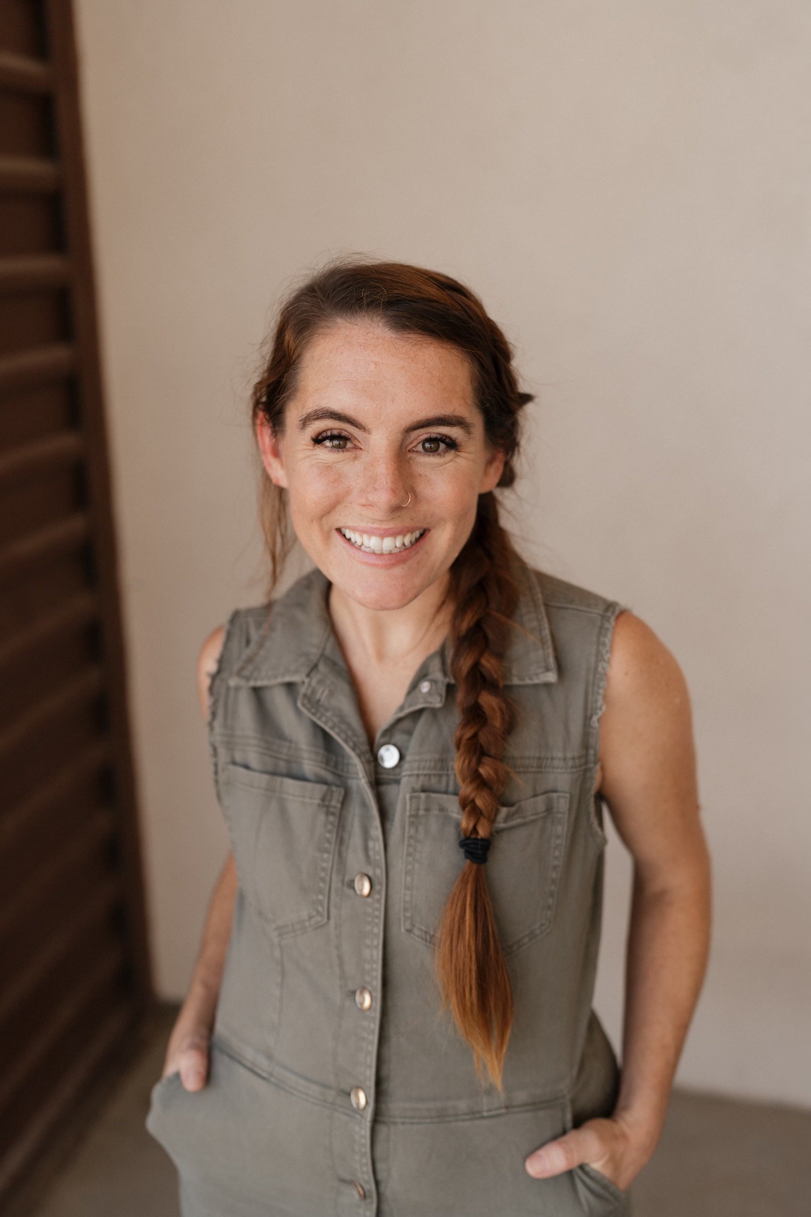 Smiling woman with a long braid, wearing a sleeveless denim vest, standing indoors against a plain wall.