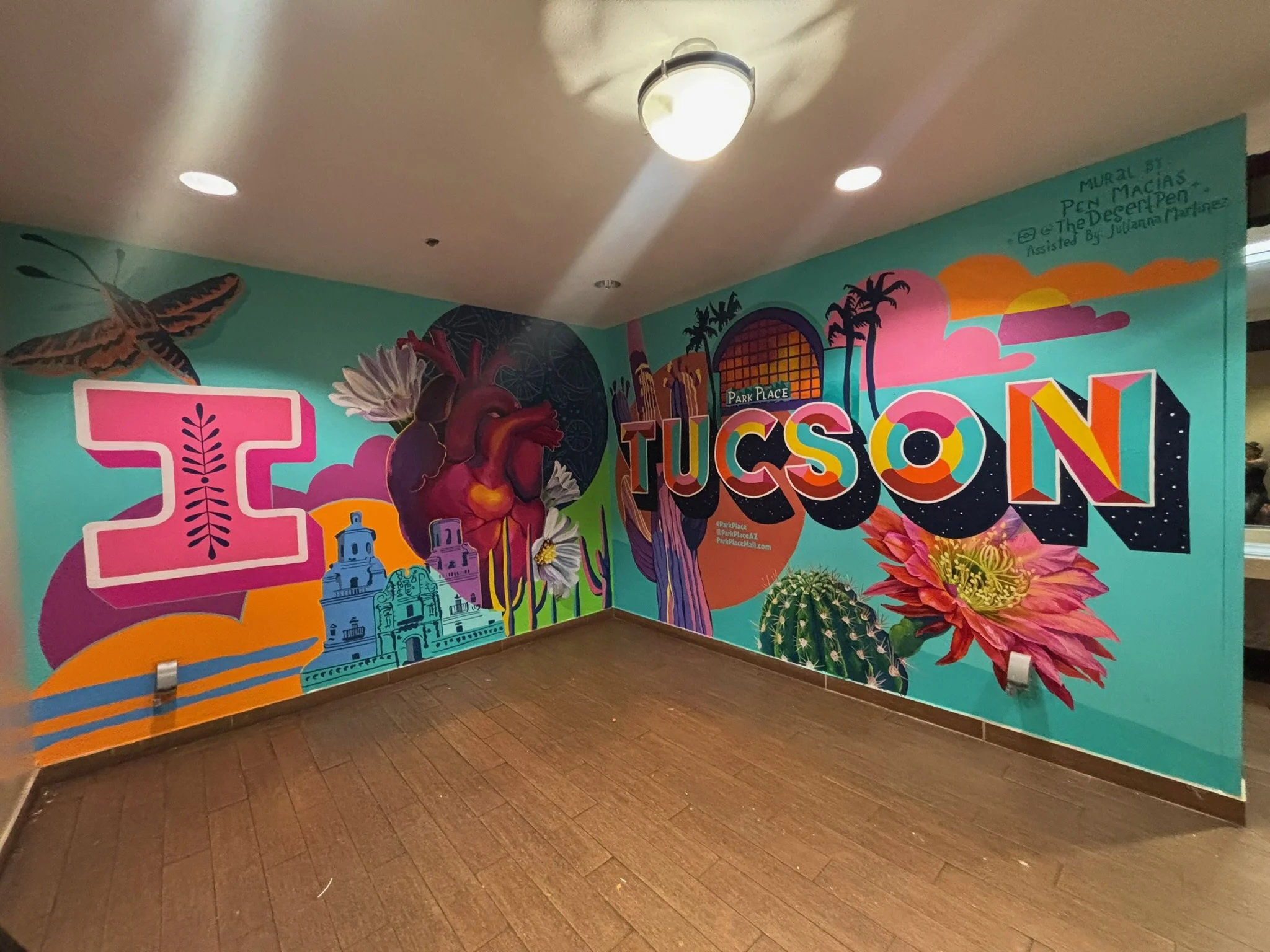 Colorful mural on a wall featuring a butterfly, a human heart with flowers, and stylized text that reads 'TUCSON' with vibrant sunset-inspired colors and images related to Tucson, Arizona.