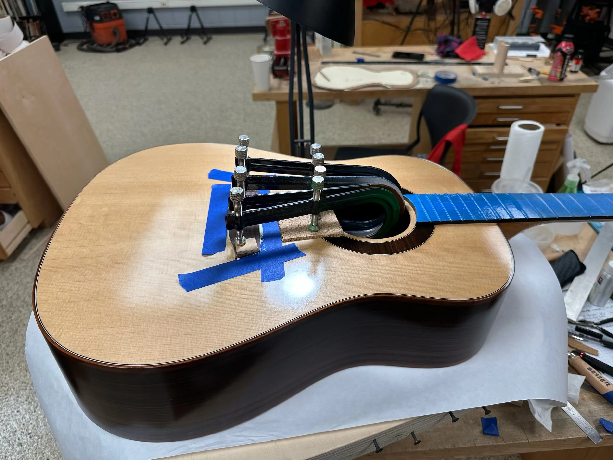 Guitar body on work table in a workshop, with clamps and masking tape holding the neck in place, surrounded by woodworking tools and materials.