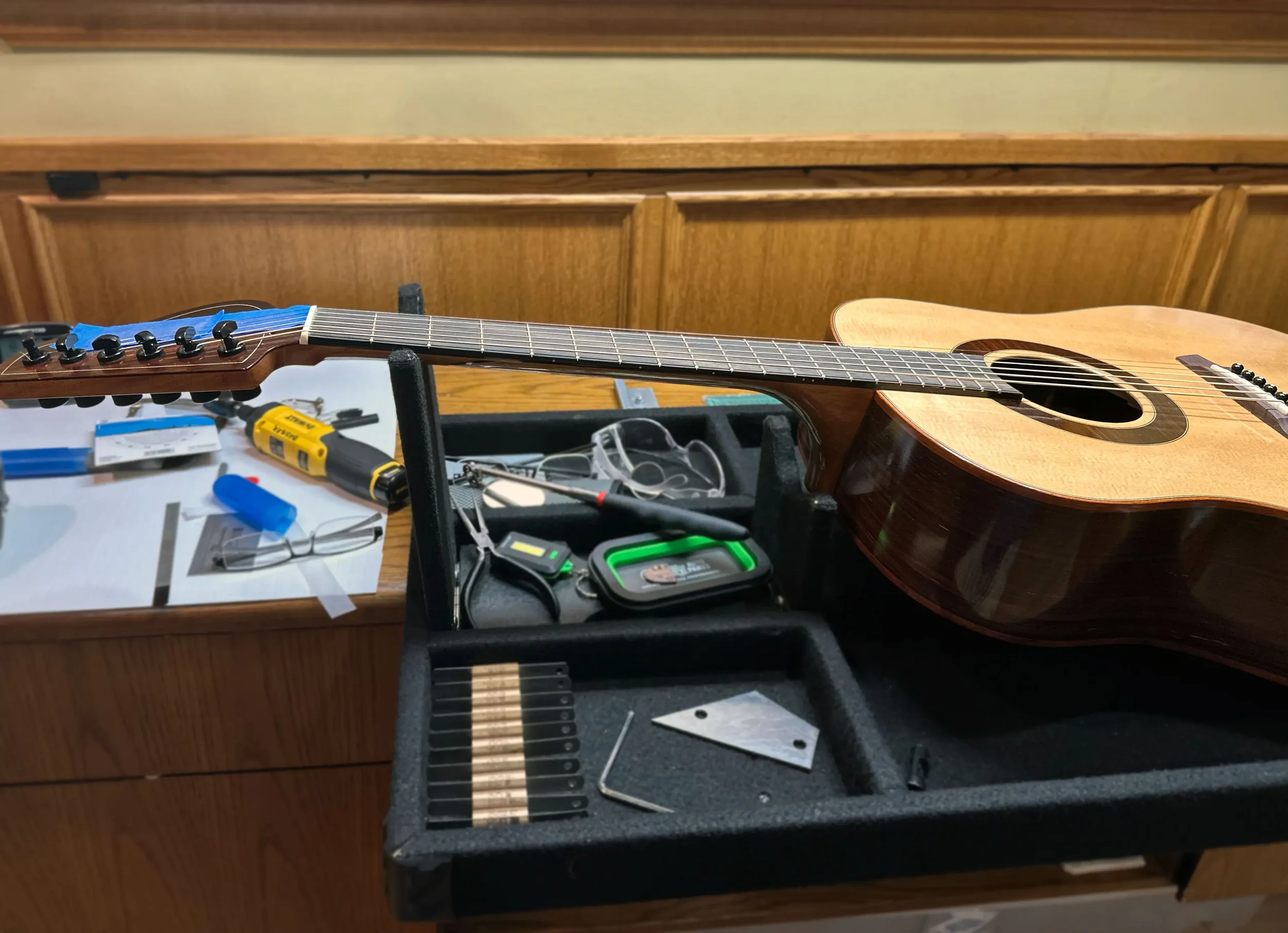 An acoustic guitar resting on a black stand, with various tools and parts around it, on a workbench in a woodworking shop.