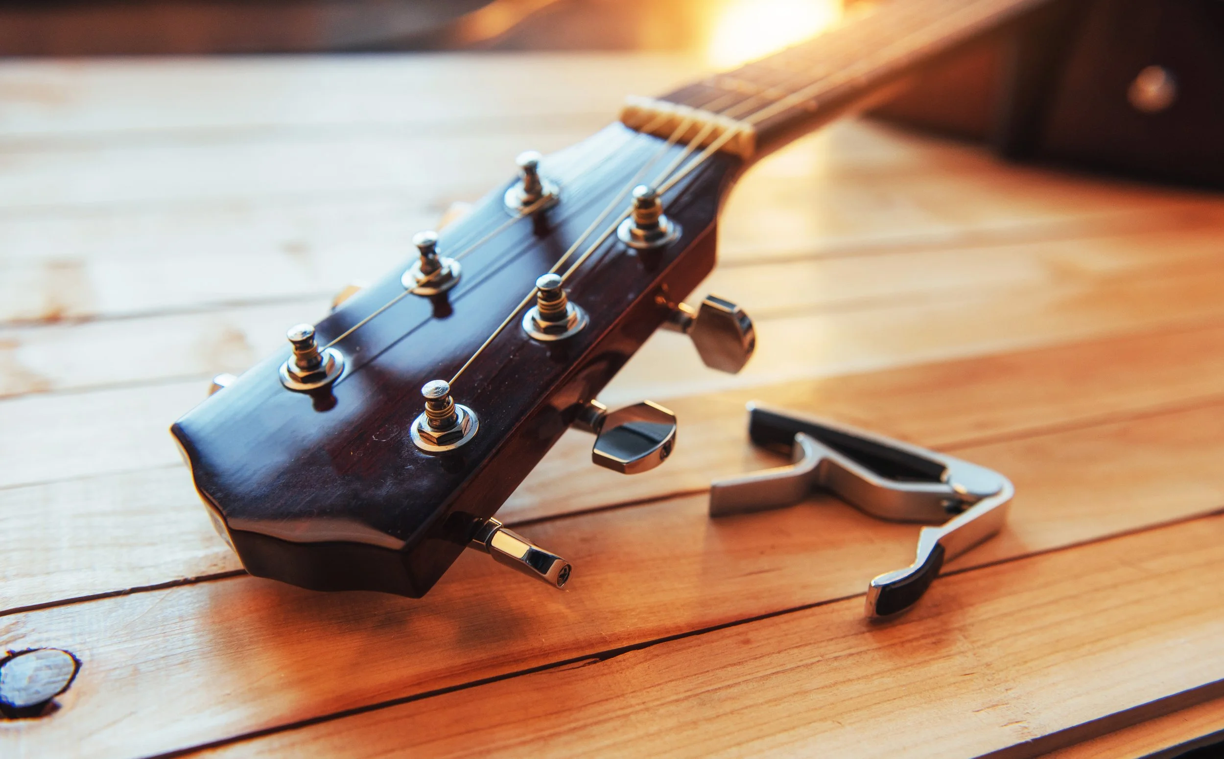 Close-up of a guitar headstock resting on a wooden surface with tuning pegs and a capo nearby.