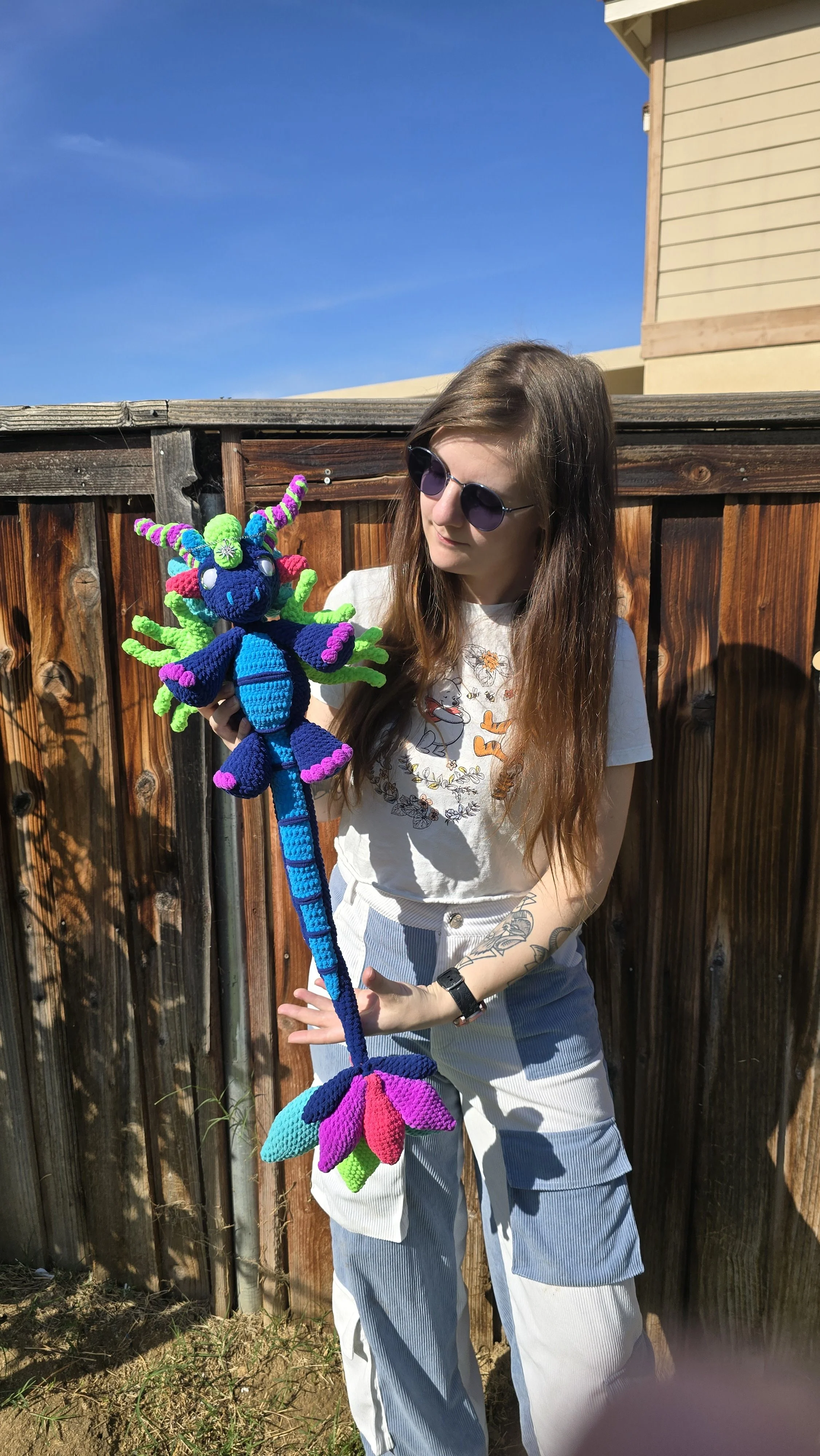 A young woman wearing sunglasses and a white T-shirt with animated characters is holding a colorful, knitted stuffed toy resembling a dragon or mythical creature, standing outdoors near a wooden fence with a clear blue sky in the background.