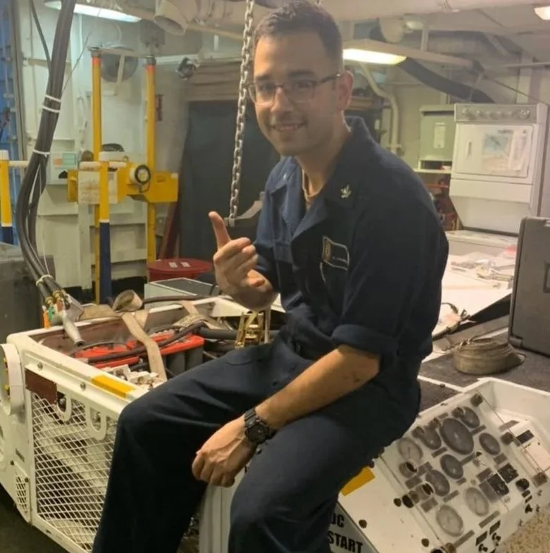 A man in a naval uniform sitting on a piece of industrial equipment in a technical or mechanical environment, smiling and making a shaka sign with his right hand.