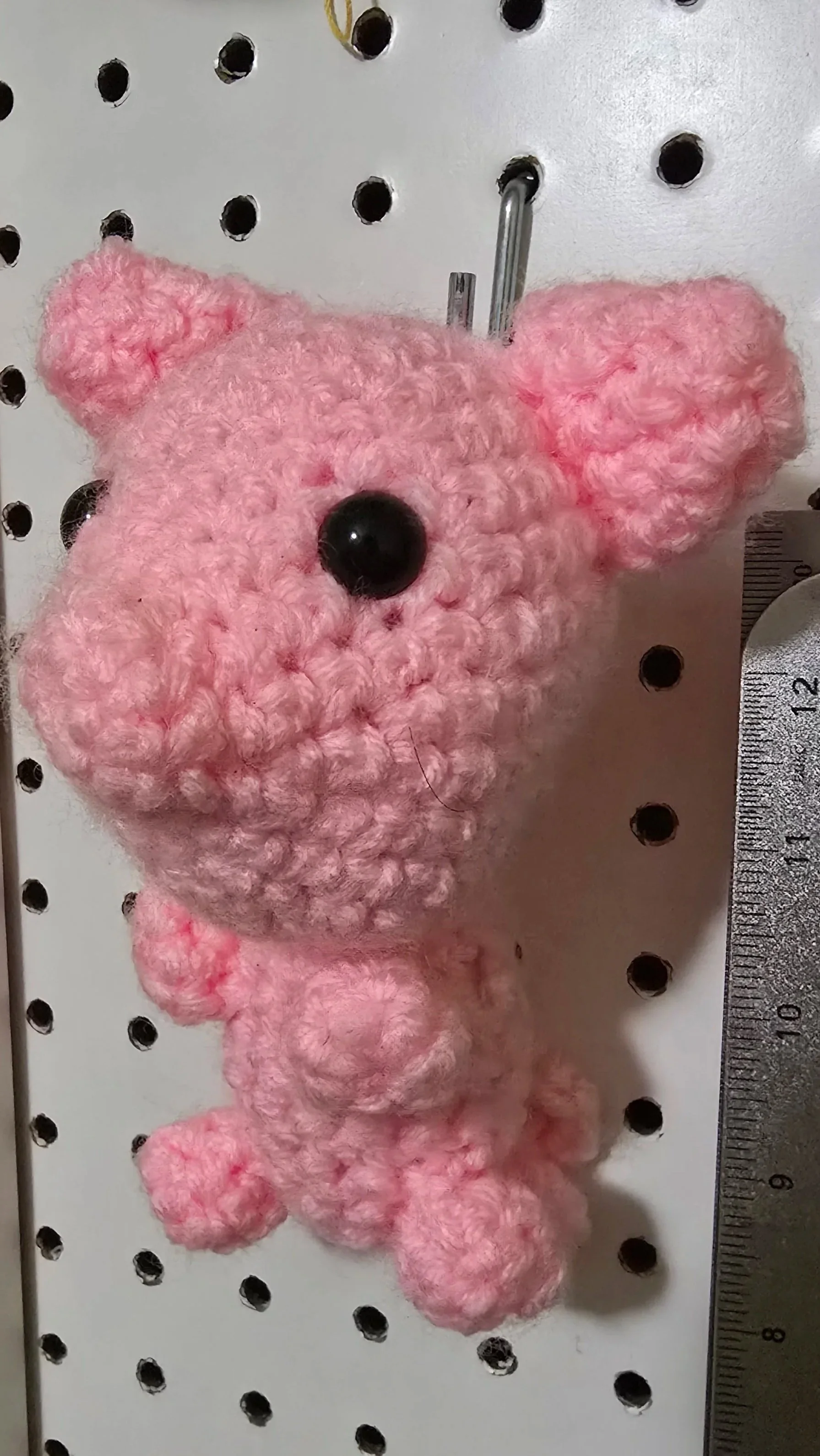 A pink, fluffy crocheted teddy bear with black button eyes and a black nose, hanging on a white pegboard with circular holes and a metal ruler at the side.