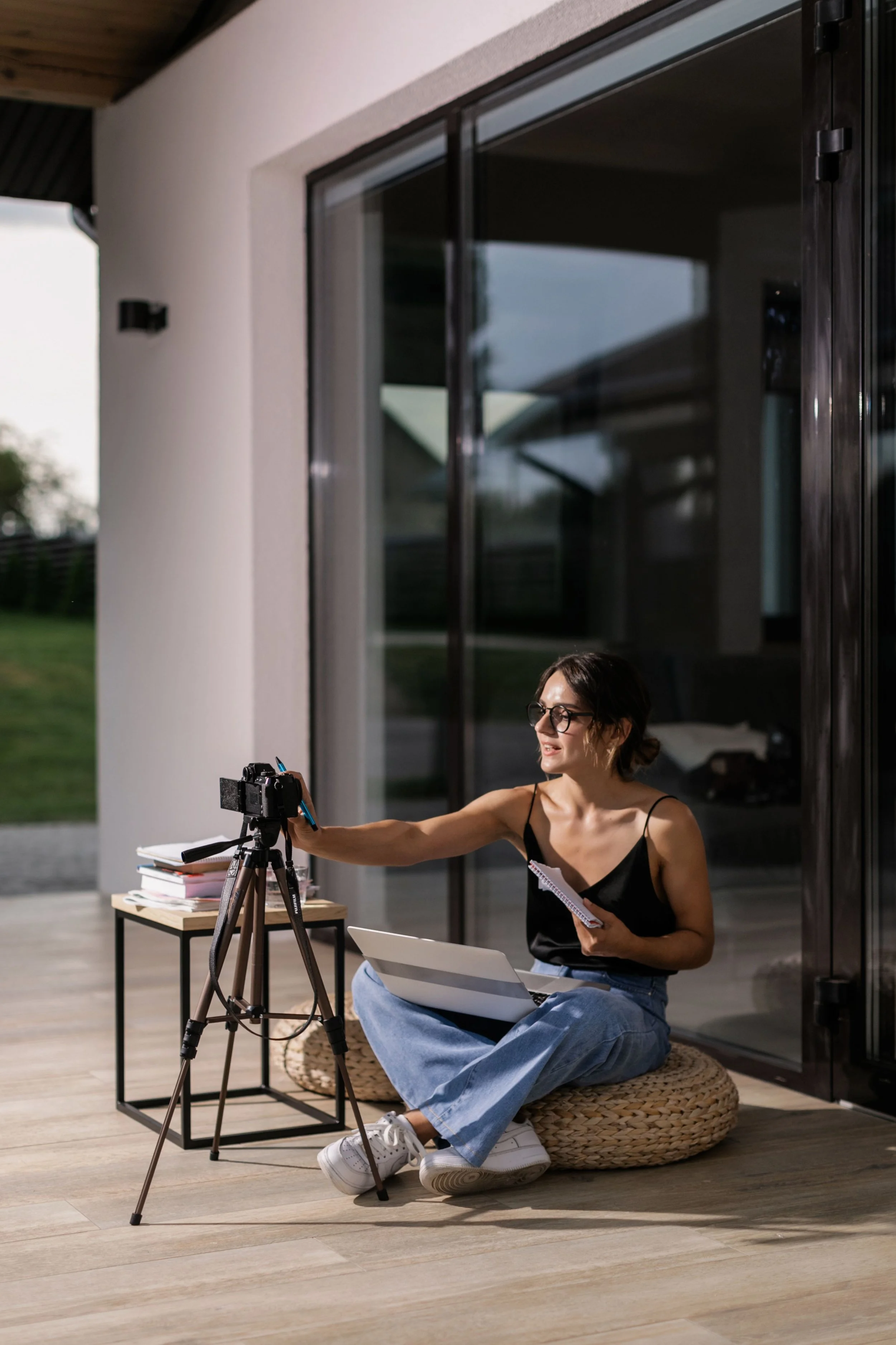 Woman sitting on floor using camera on tripod, holding notepad, in a room with large sliding glass doors and a small table with books and notebooks.