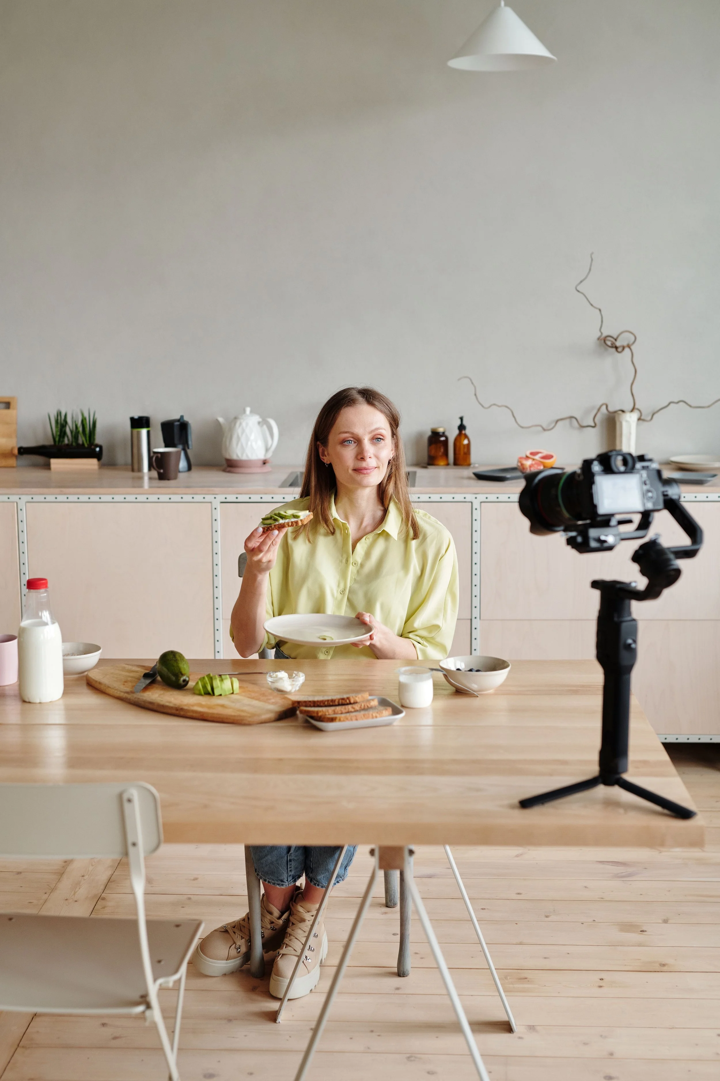 A woman wearing a yellow shirt and jeans shorts is holding a plate and a slice of cucumber in a kitchen, with a camera on a tripod in front of her, and various kitchen items and ingredients on the wooden table.