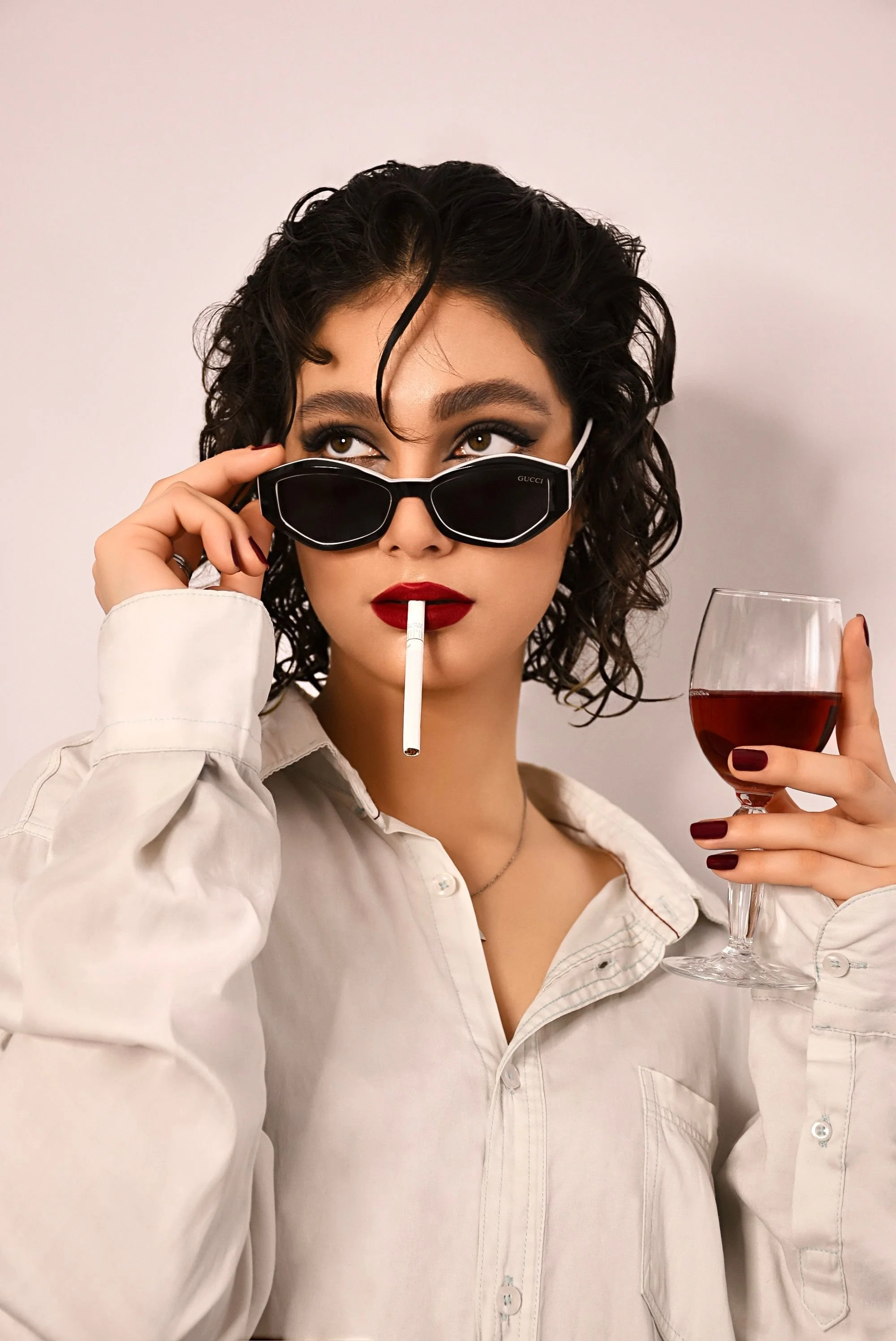 A woman with curly dark hair, wearing a white button-up shirt, black Gucci sunglasses, and red lipstick, holding a glass of red wine in one hand and a cigarette in her mouth, while adjusting her sunglasses with her other hand.