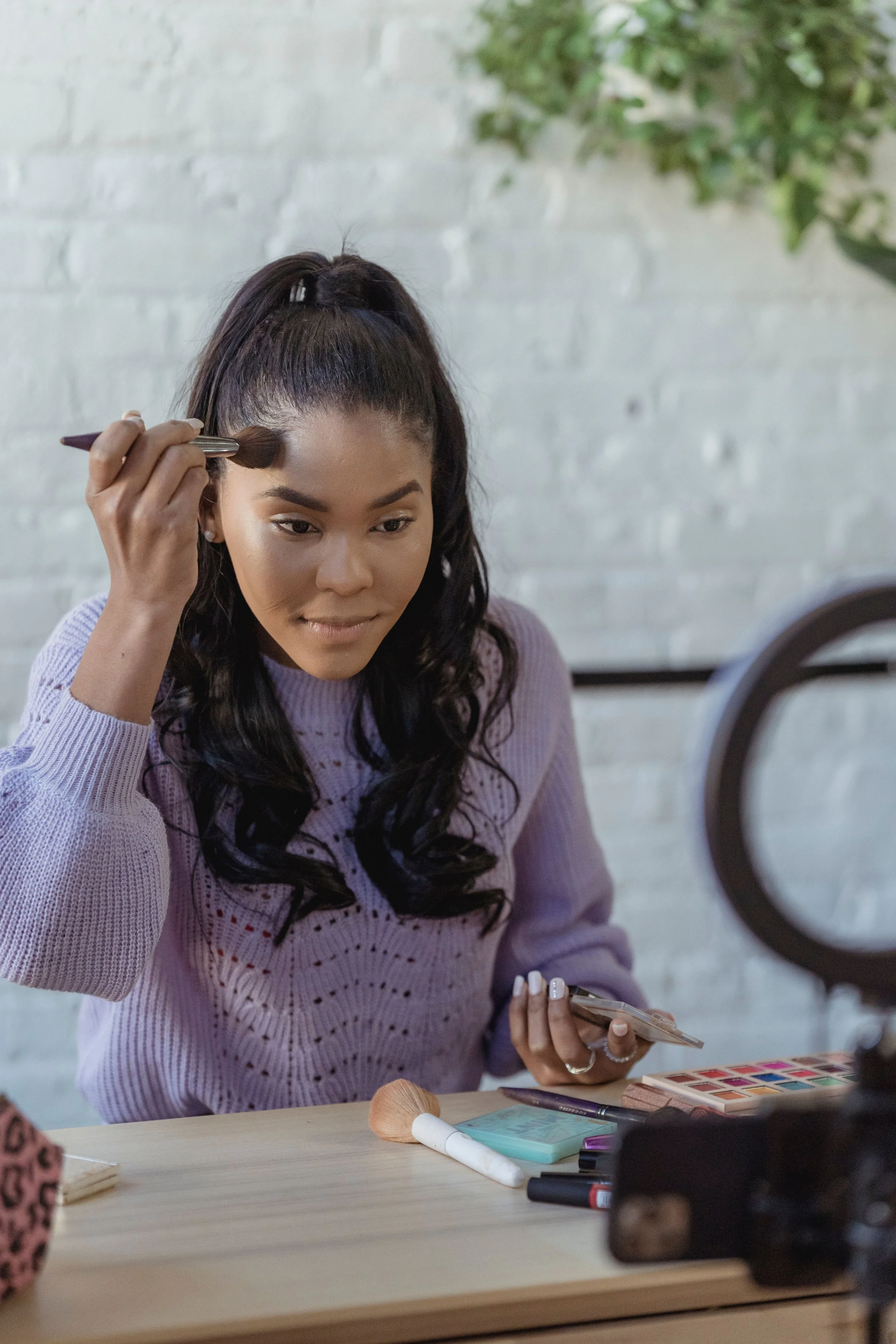 A woman is sitting at a table, applying makeup with a brush, holding a makeup palette and a phone, with makeup tools and products on the table. She is wearing a lavender sweater, has dark wavy hair, and appears focused on her makeup routine.