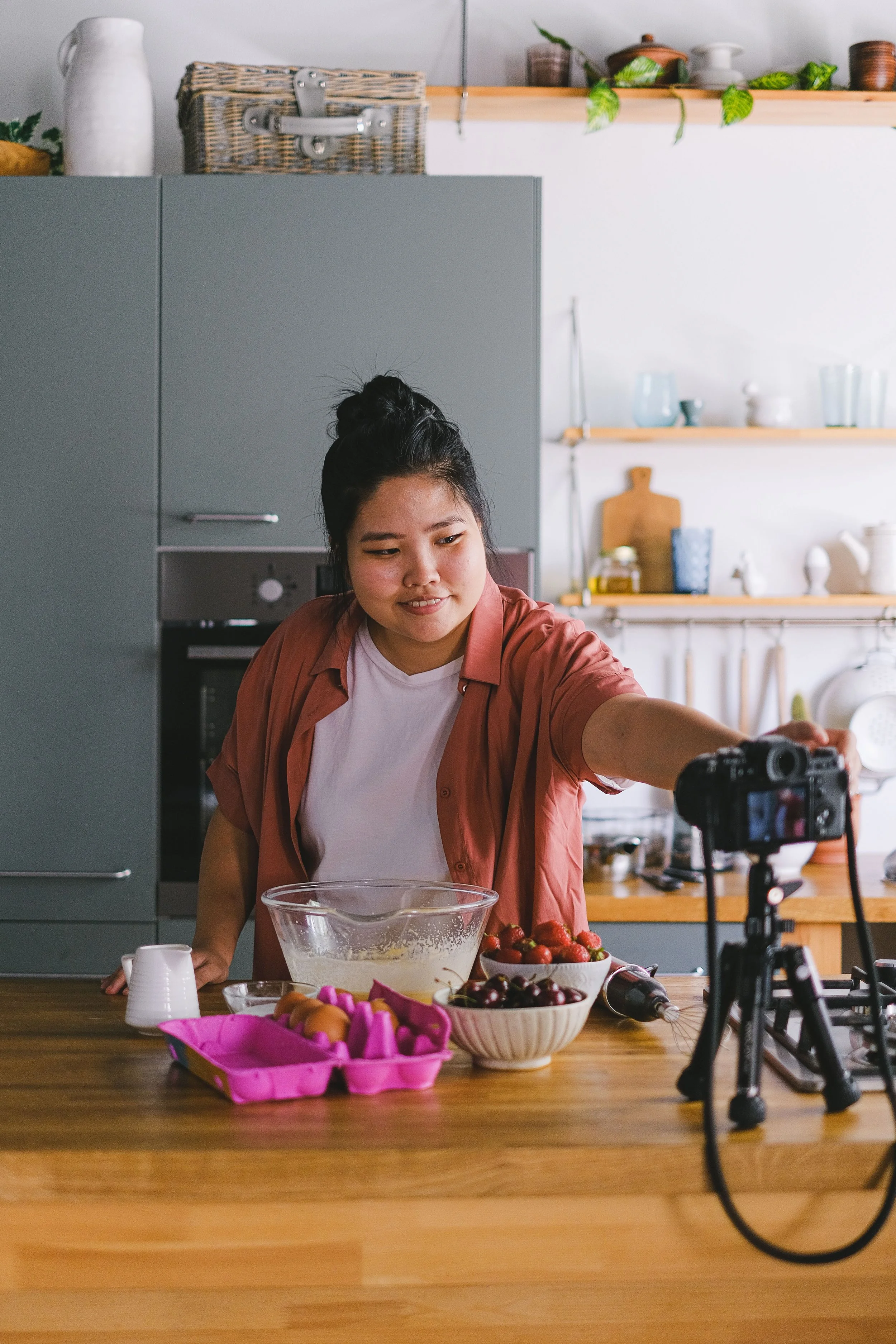 A young woman with dark hair in a bun is in a kitchen, preparing food. She is leaning over a table that has bowls of strawberries and other produce, a pink egg carton with eggs, and a clear mixing bowl. She is wearing a white T-shirt under an open salmon-colored shirt. There is a camera on a tripod set up on the table, recording her. The background has grey cabinets and open wooden shelves with various kitchen items.