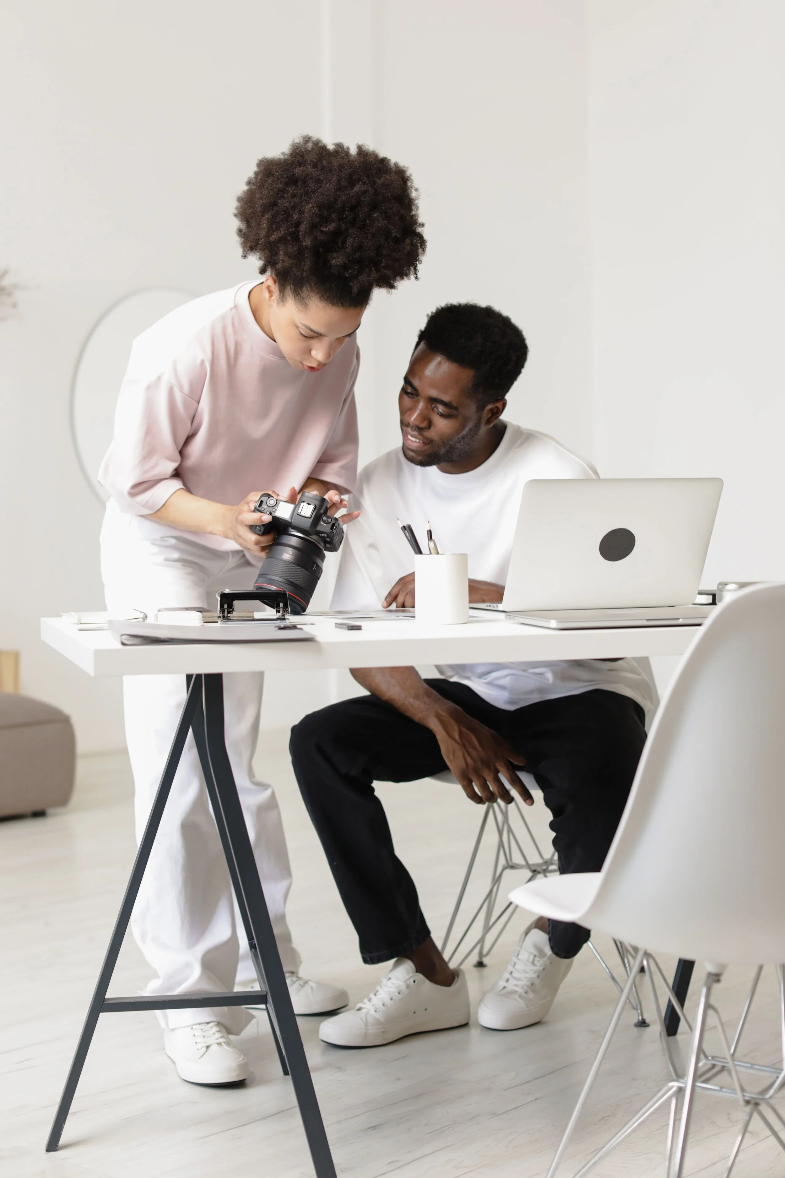 Two young adults, a woman and a man, are working together at a white desk in a bright, minimalist office. The woman, with curly hair, is standing and showing something on a camera to the seated man, who is looking at her with interest. The man has short, dark hair and is wearing a white t-shirt. There is a laptop, a white pen holder with pens, and some papers on the desk.