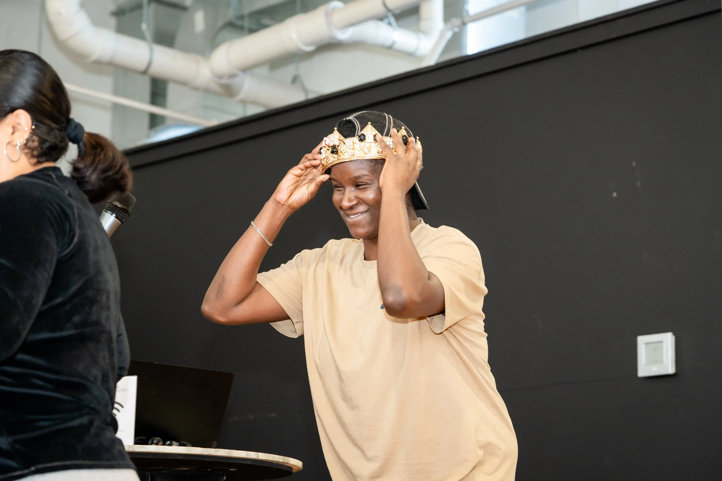 A young woman is smiling and adjusting a crown on her head while standing next to a woman holding a microphone. The background shows an industrial-style setting with pipes and a black wall.