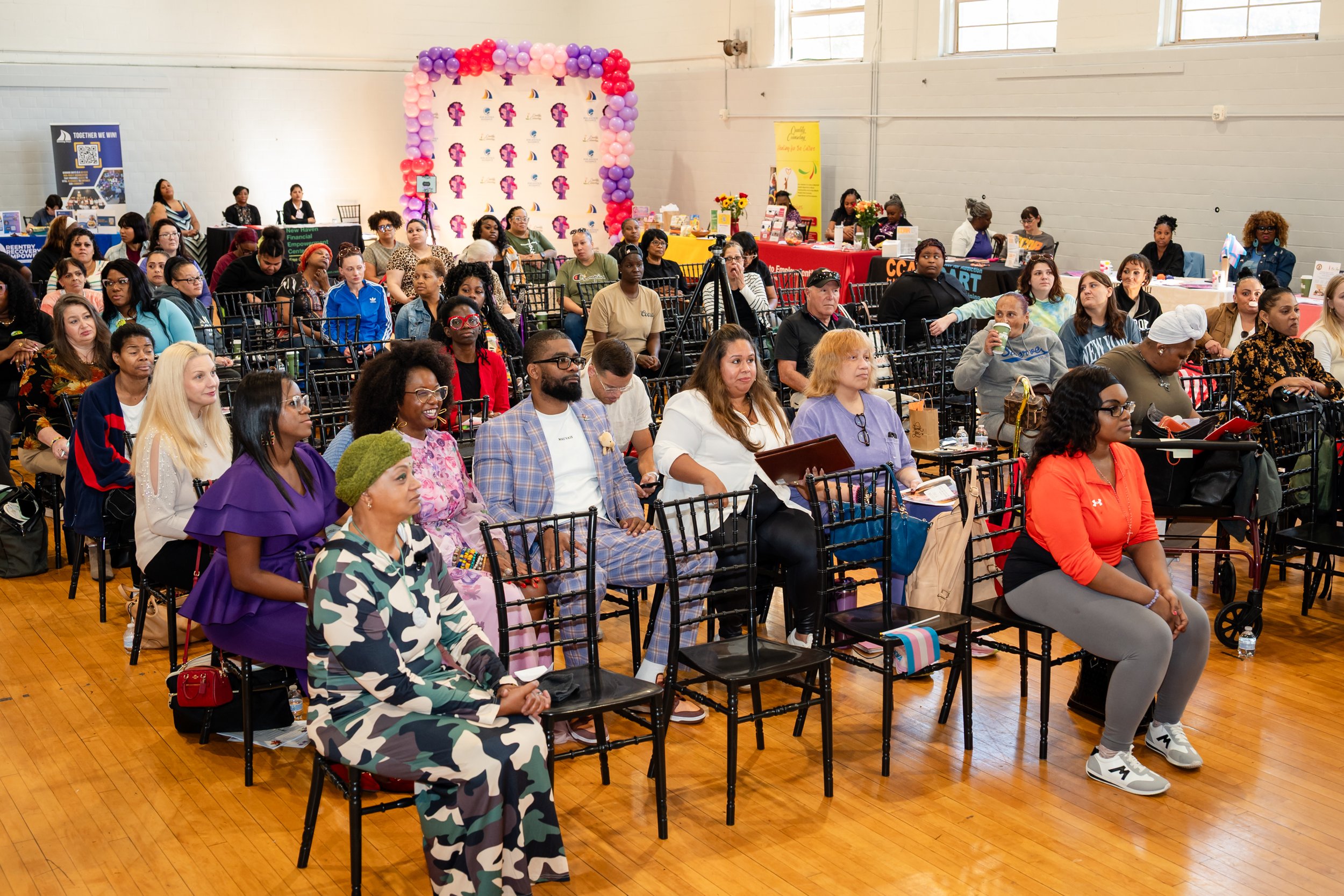 A diverse group of women and a few men seated in folding chairs in a large indoor space attending an event. There are tables in the background with banners, balloons, and informational displays.