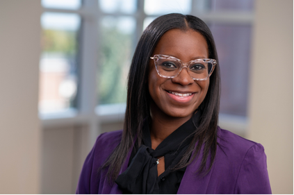 A woman smiling, wearing glasses, a purple blazer, and a black scarf in an office setting.