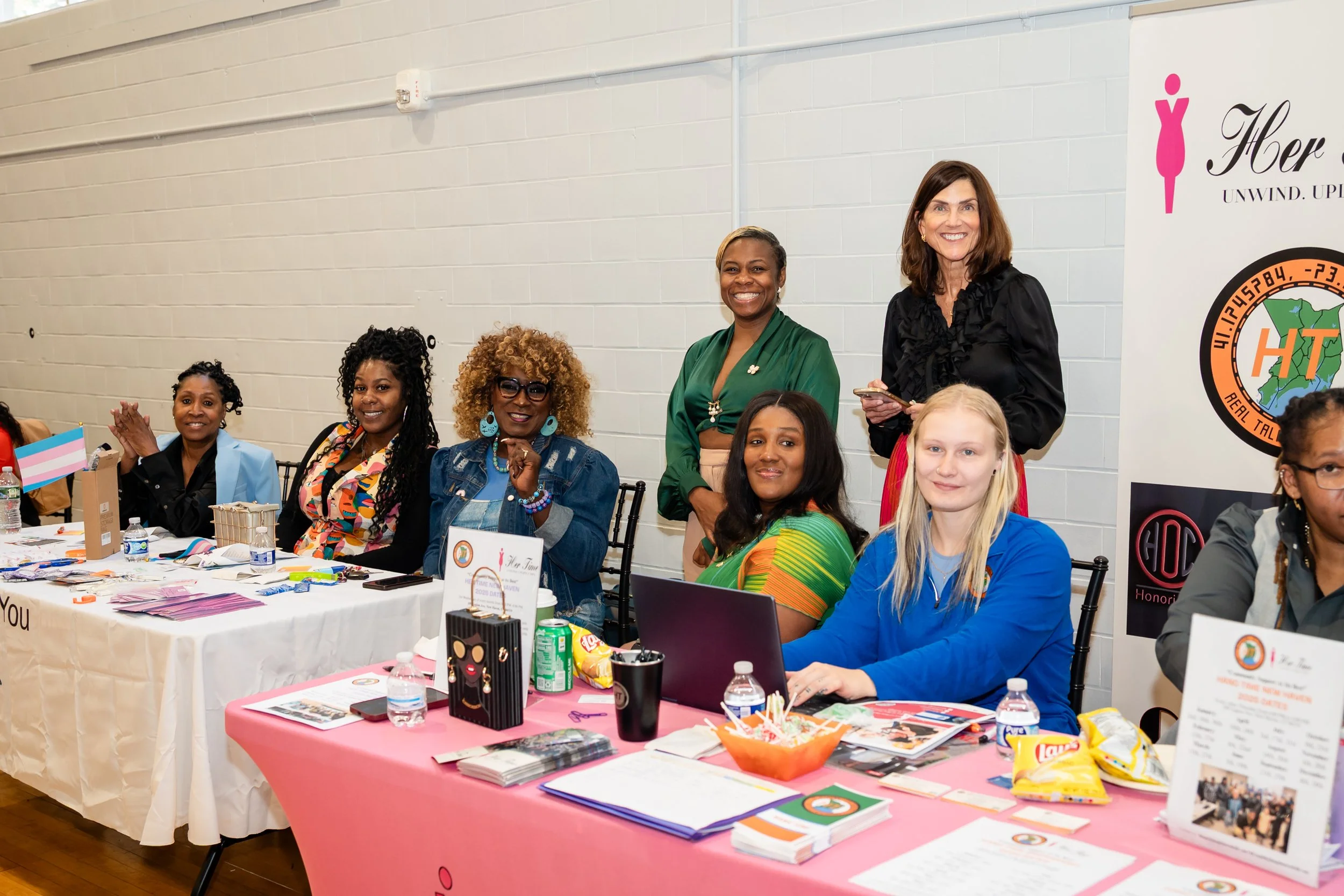 A group of women sitting at tables during an indoor event, smiling at the camera, with various informational and promotional materials on the tables.