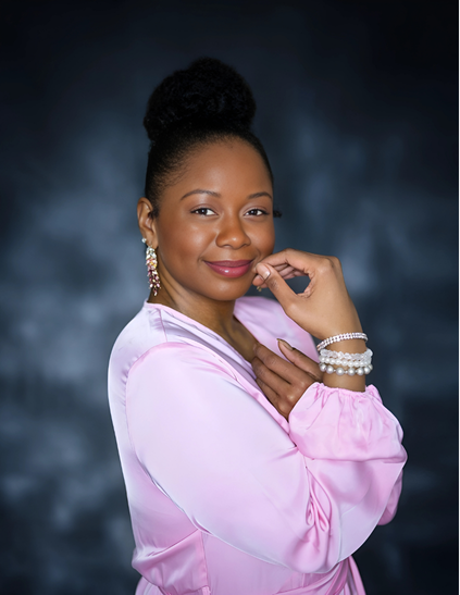 Portrait of a woman with dark hair styled in a bun, wearing a pink satin blouse and pearl jewelry, smiling softly against a dark background.