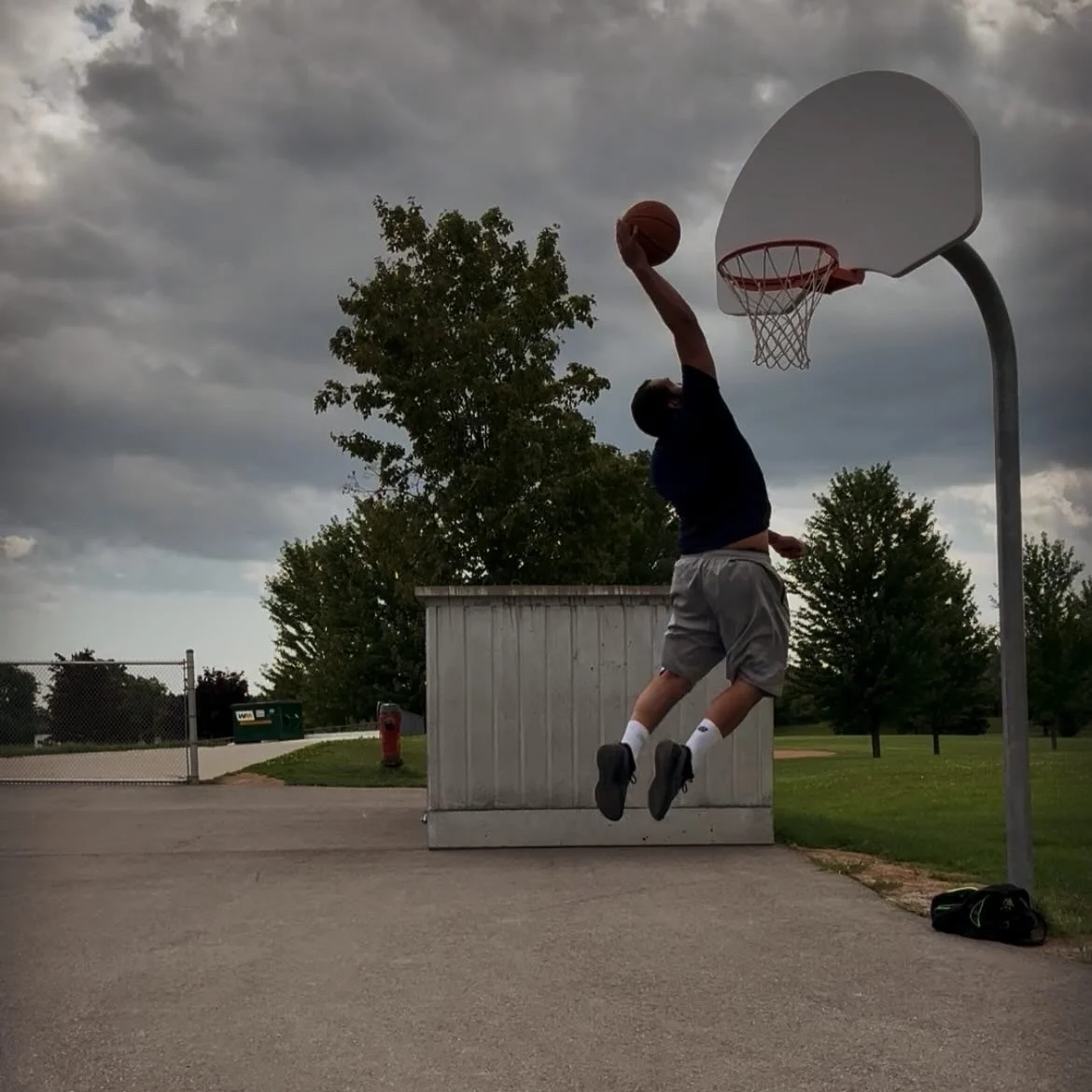 A person jumping in mid-air while shooting a basketball near a basketball hoop outdoors on a cloudy day.