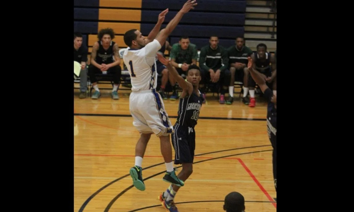 A basketball game with players on the court, one player in white jumping to block a shot from a player in black, while other players and spectators Bench sit in the background.