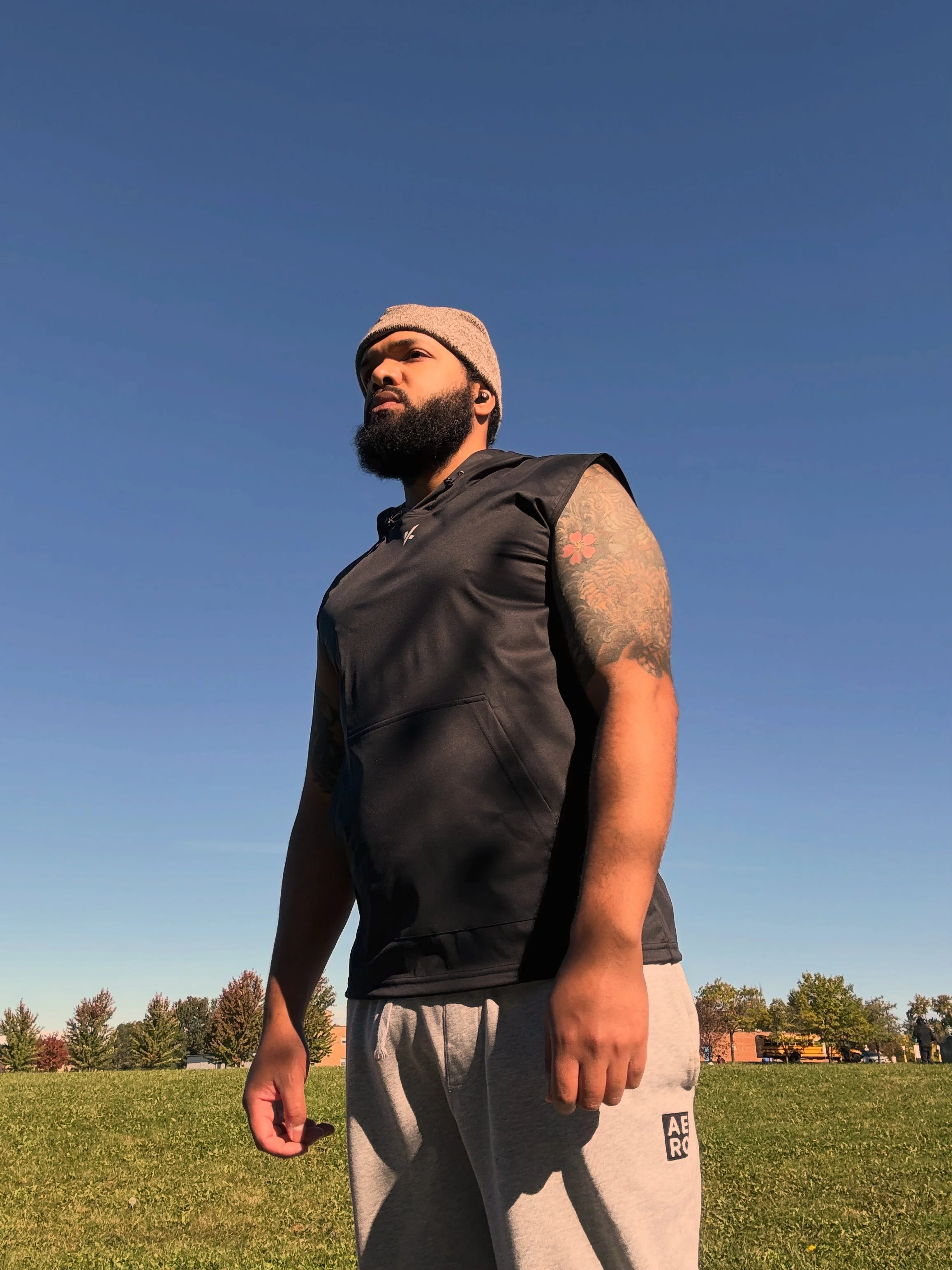 A man with a beard and tattoos wearing a beige beanie, black sleeveless shirt, and gray shorts stands outdoors on grass with trees and houses in the background, under a clear blue sky.
