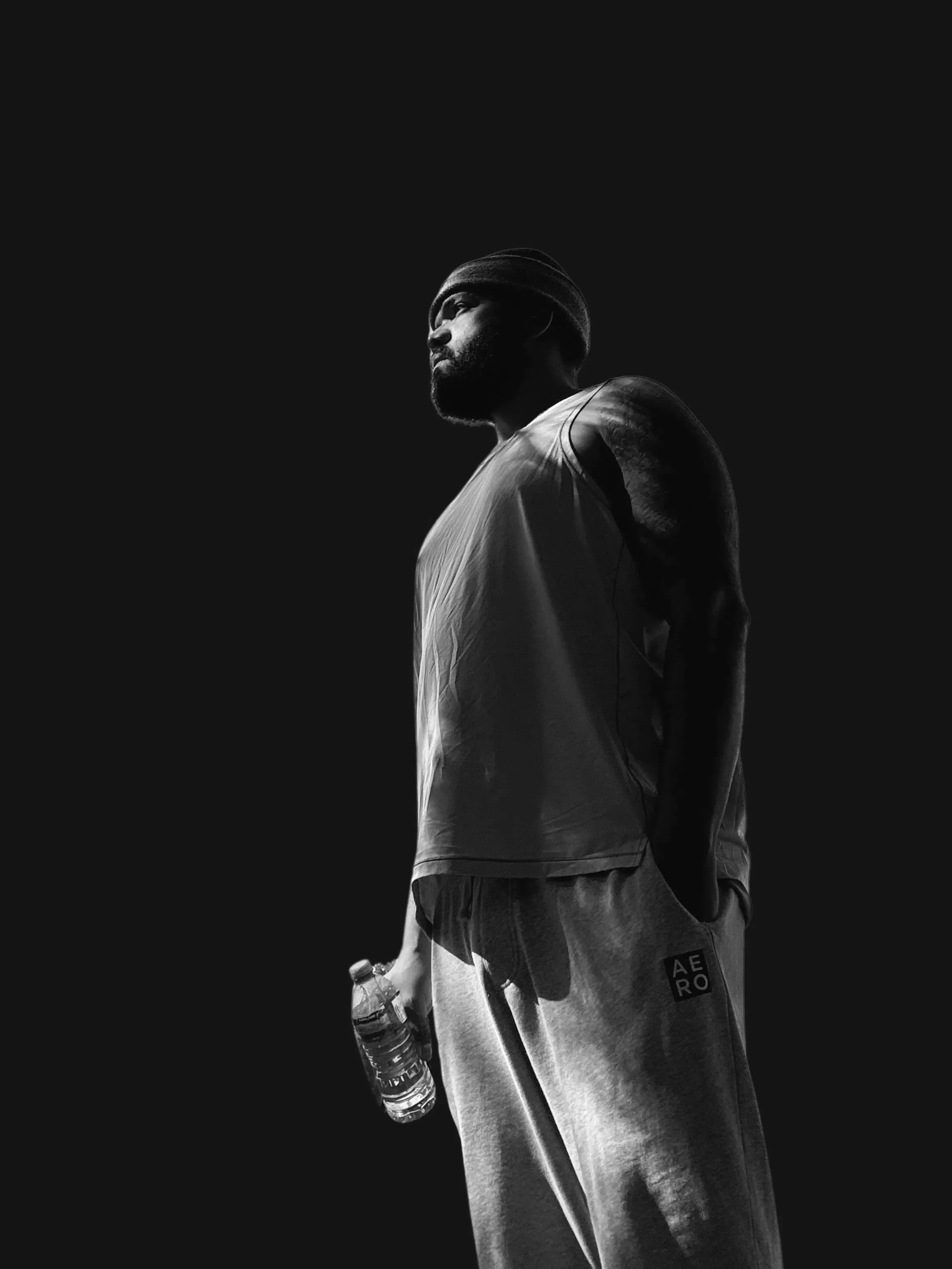 A black and white photo of a muscular man wearing a beanie, tank top, and sweatpants, holding a water bottle, standing against a dark background.