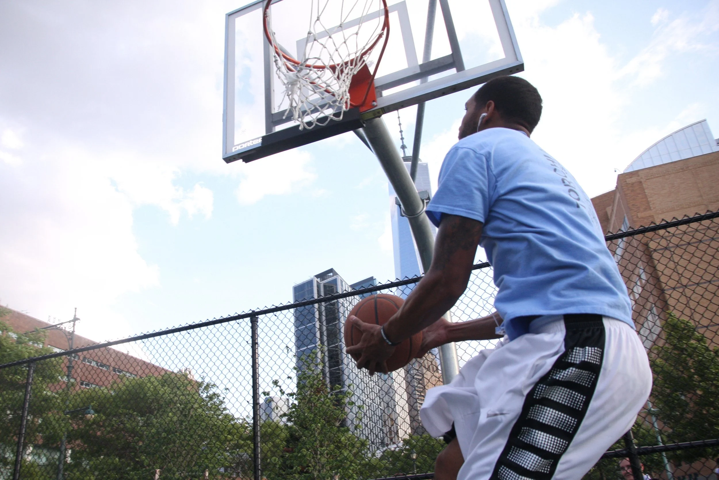 A man in a blue shirt holding a basketball, standing on an outdoor basketball court with a basketball hoop and chain-link fence, city buildings in the background.