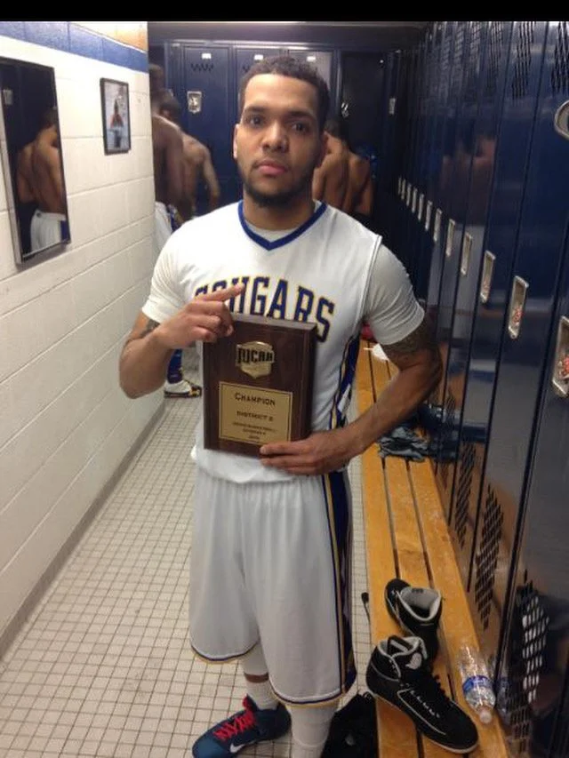 A young male basketball player standing in a locker room, holding a plaque that says 'Champion,’ wearing a white jersey with blue and yellow accents and the word 'Organigas' on it.