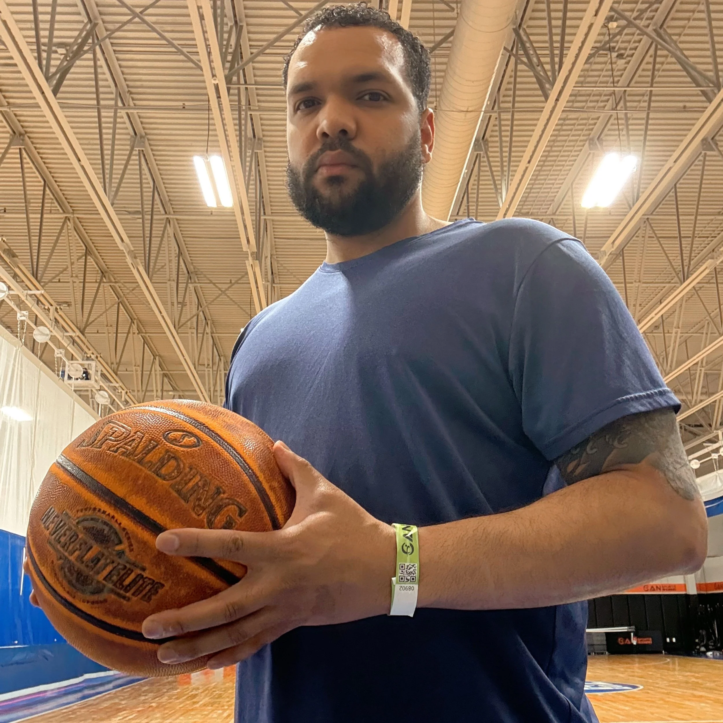 Man with dark curly hair and beard, wearing a dark shirt, holding a basketball, in a gymnasium with a high ceiling, metal framework, bright lighting, and a basketball court in the background.