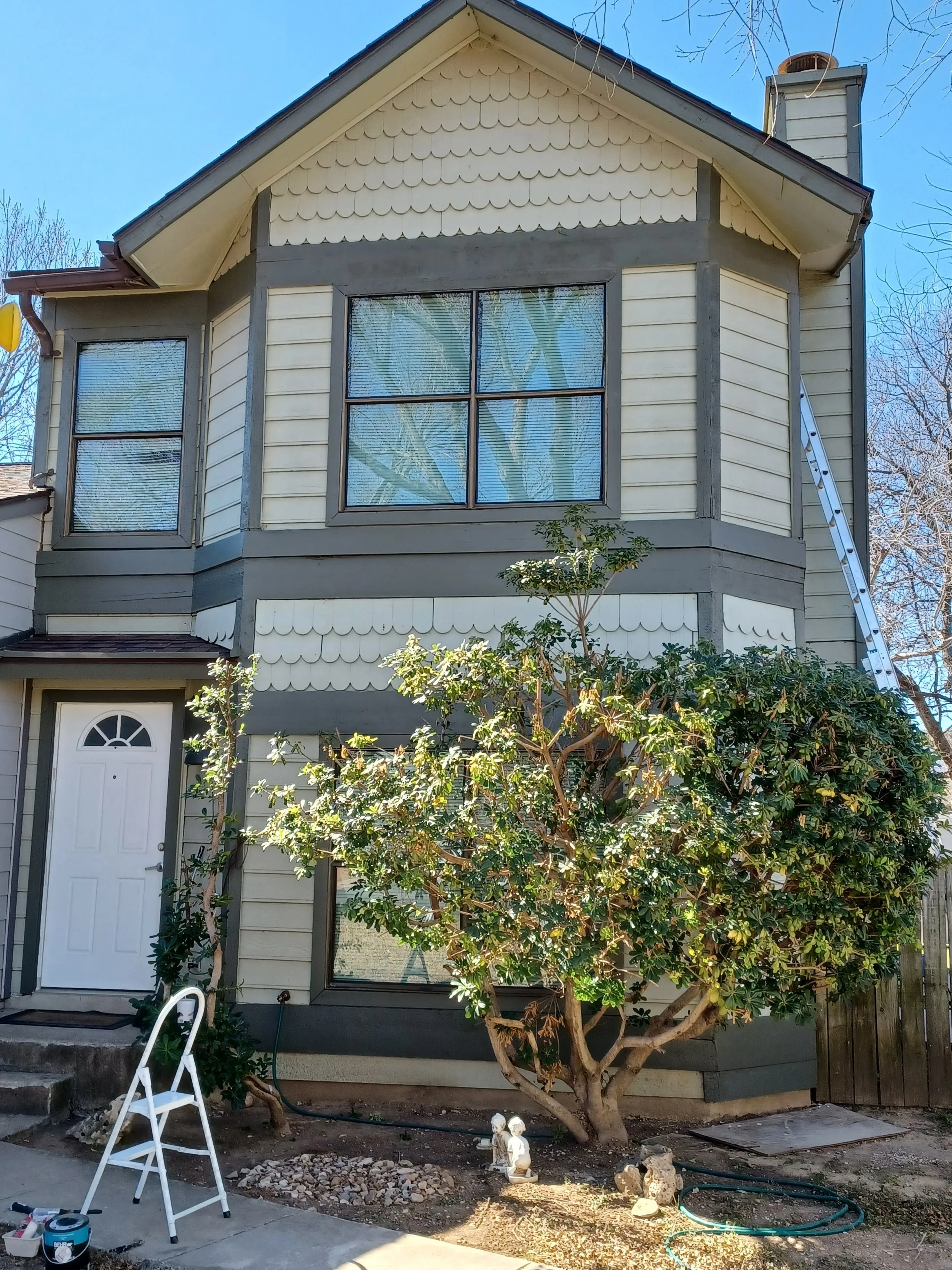 Front view of a house with a bay window on the second floor, a white front door, and a small front yard with a tree and some garden decorations. There is a tall ladder leaning against the house and some gardening tools and materials on the ground.