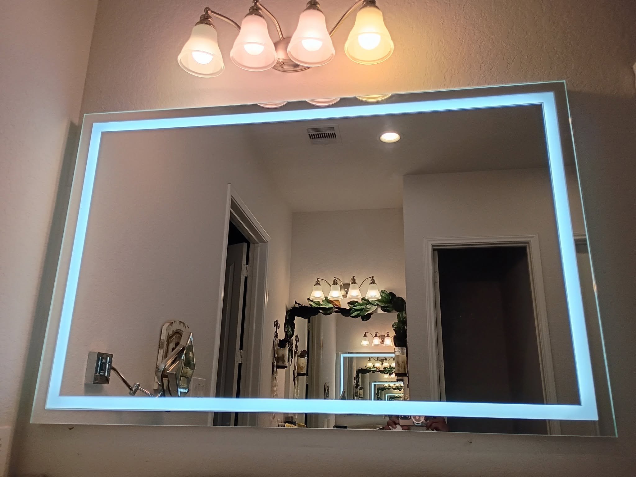 A bathroom mirror with LED lighting around the frame, showing the reflection of a room with multiple ceiling lights, wall sconces, and various plants and decorative items.